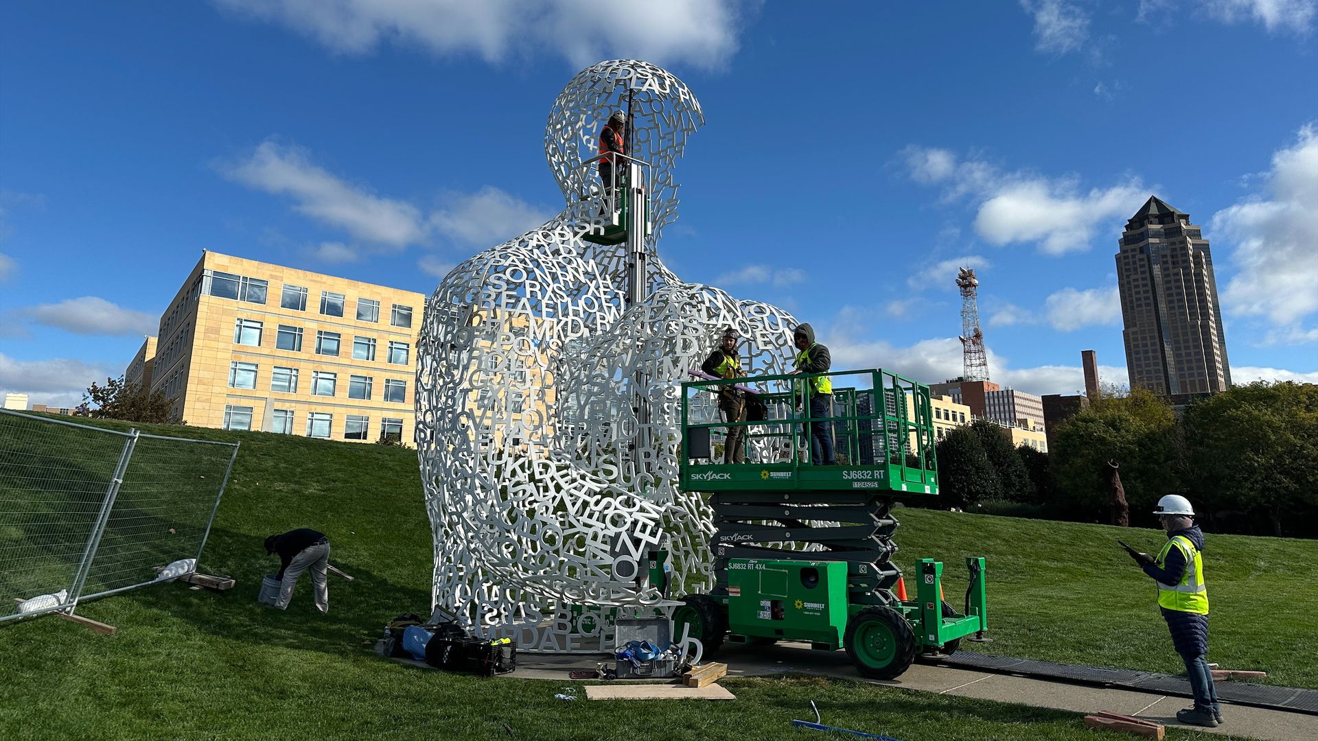 Workers in yellow safety vests and helmets install a rehabilitate Nomade, a tall white sculpture made of interlocking letters in an outdoor park with green grass, blue sky, and buildings in the background.
