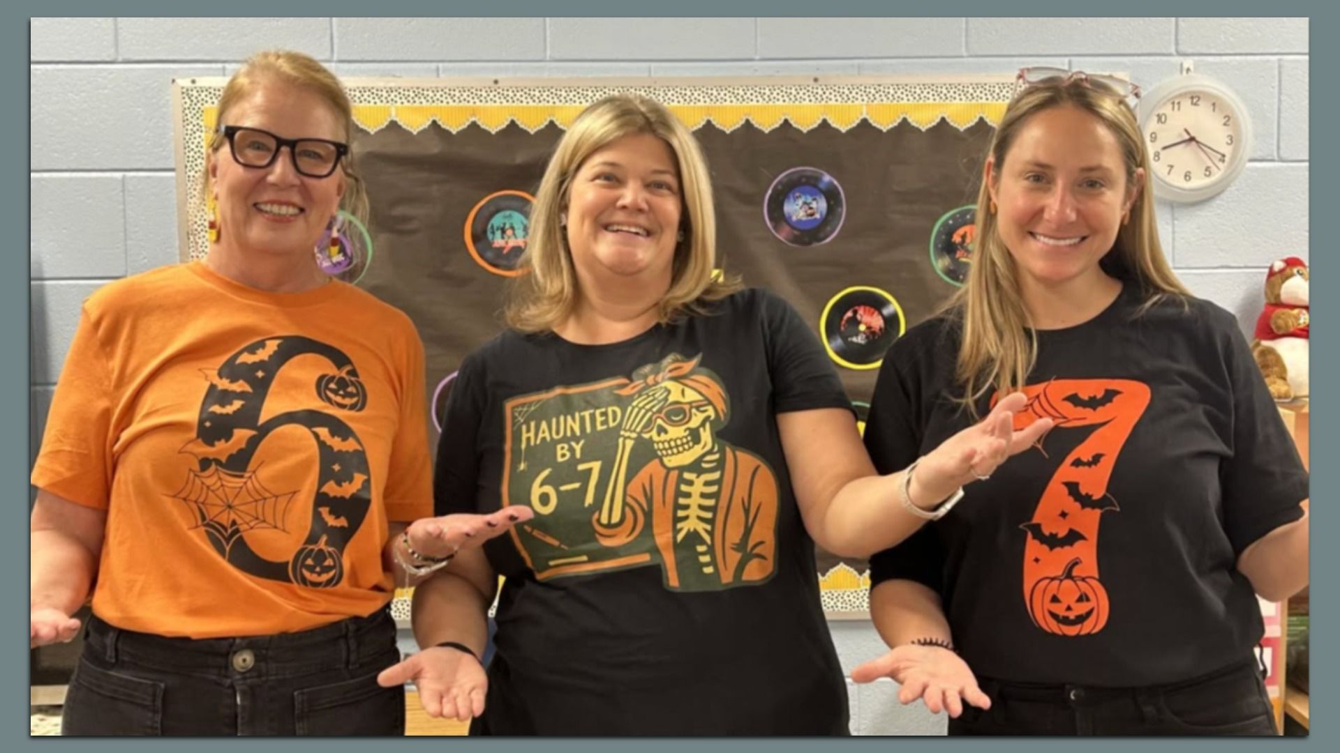 Three smiling women in a classroom wearing Halloween-themed shirts, with numbers 6 and 7 in orange and black designs, including bats, pumpkins, and skeleton illustrations.