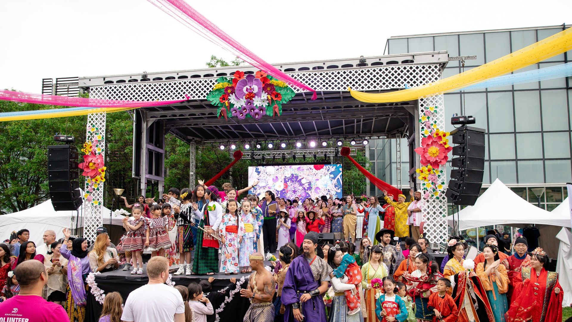Crowd of people in colorful traditional clothing on an outdoor stage decorated with bright flowers and pink and yellow streamers, with a modern glass building and trees in the background.