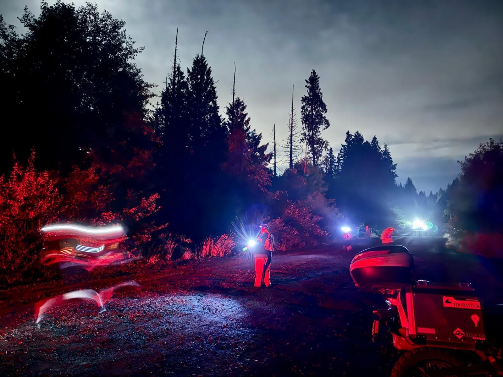 Nighttime forest road with red and blue emergency lights. A person in reflective gear holds a flashlight; blurred vehicle light trails and a boat sit to the right.