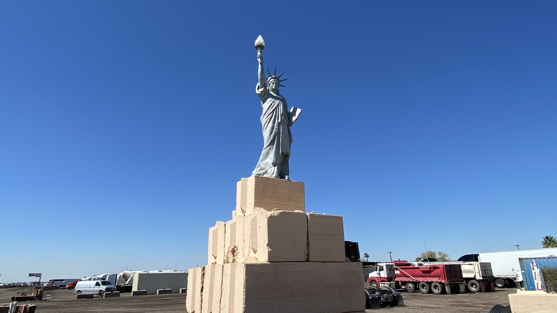 A replica of the Statue of Liberty atop concrete blocks in front of a lot filled with trucks. 