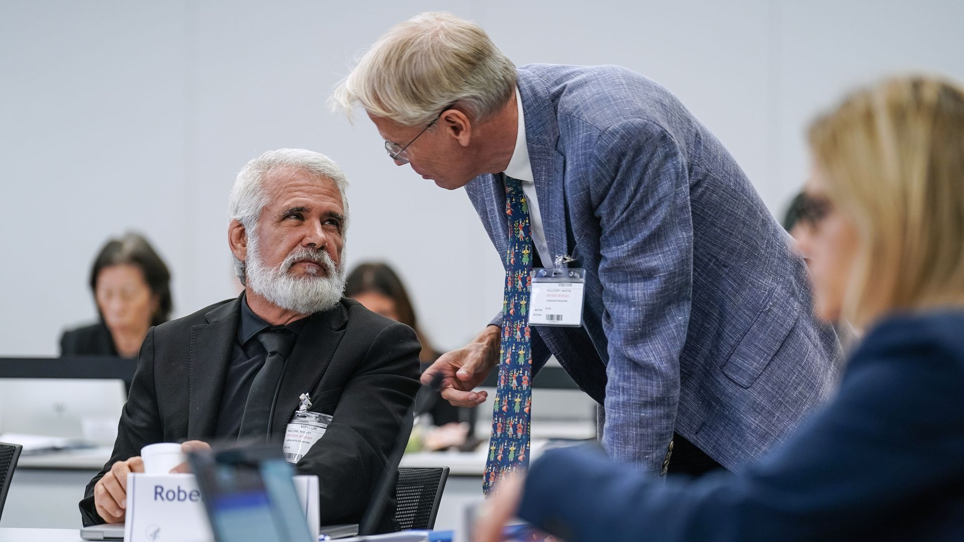 Two men in suits speak with each other. Robert Malone is sitting at a table, and Martin Kulldorf is leaning over him as the two talk.