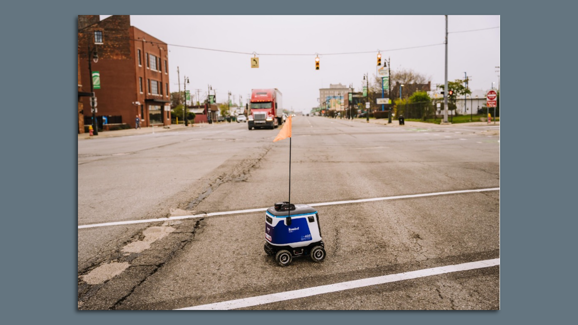 Image of a tiny delivery robot trying to cross a broad boulevard with a heavy truck approaching.