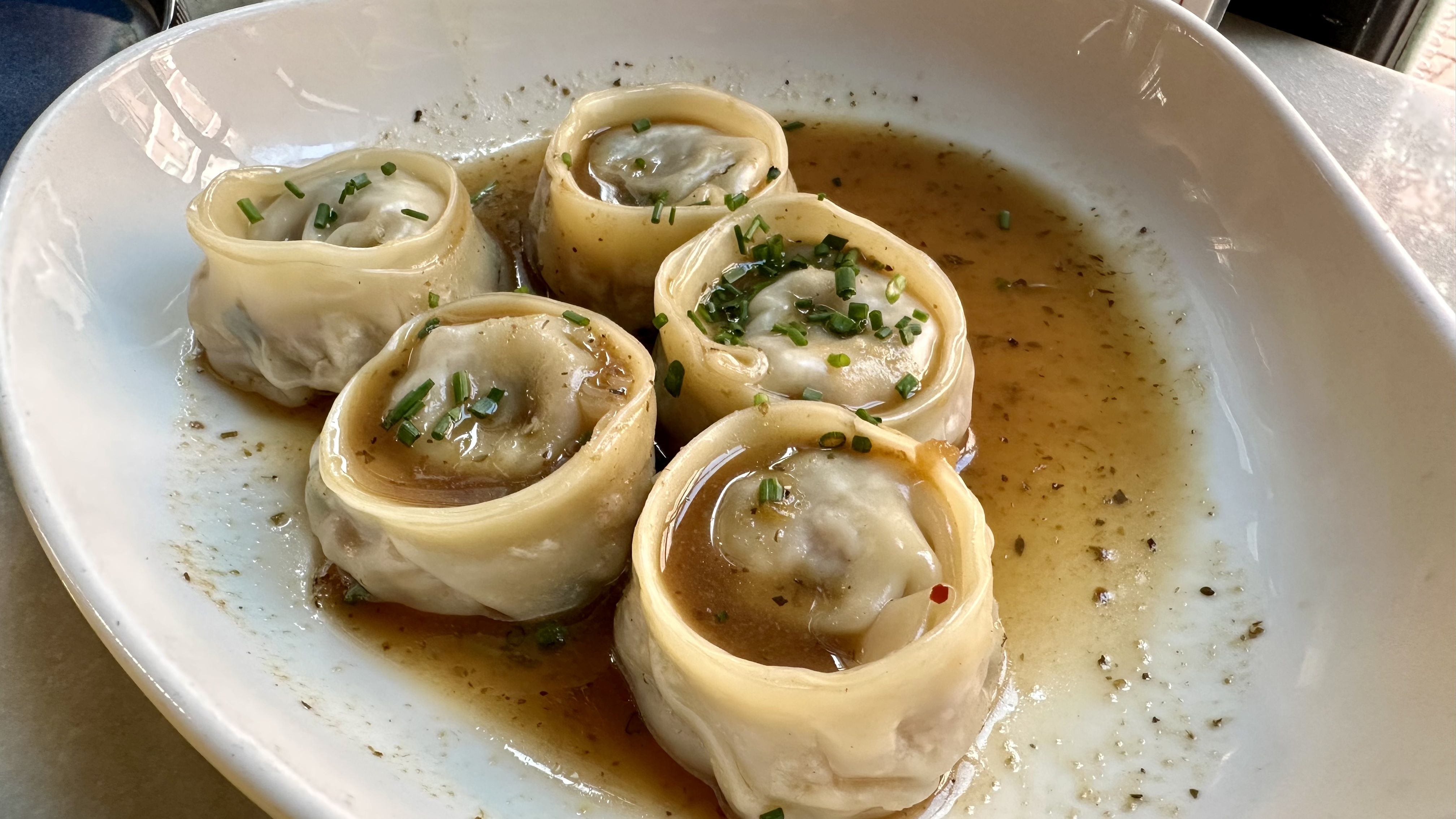 Several plump dumplings with pale wrappers stand upright in a white bowl, surrounded by light brown sauce and sprinkled with chopped chives.