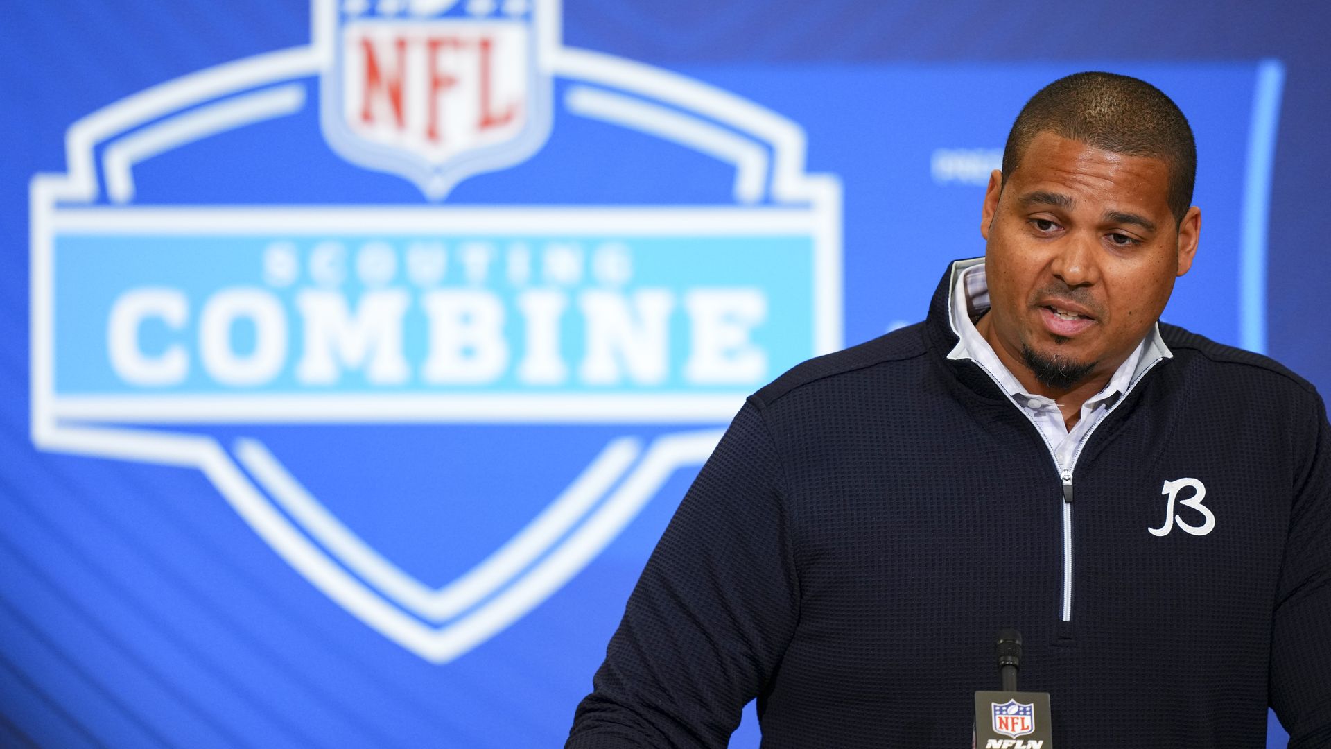 Man at a podium during an NFL Scouting Combine press conference, wearing a dark zip-up sweater with a white logo, in front of a blue backdrop featuring the NFL shield and "SCOUTING COMBINE".