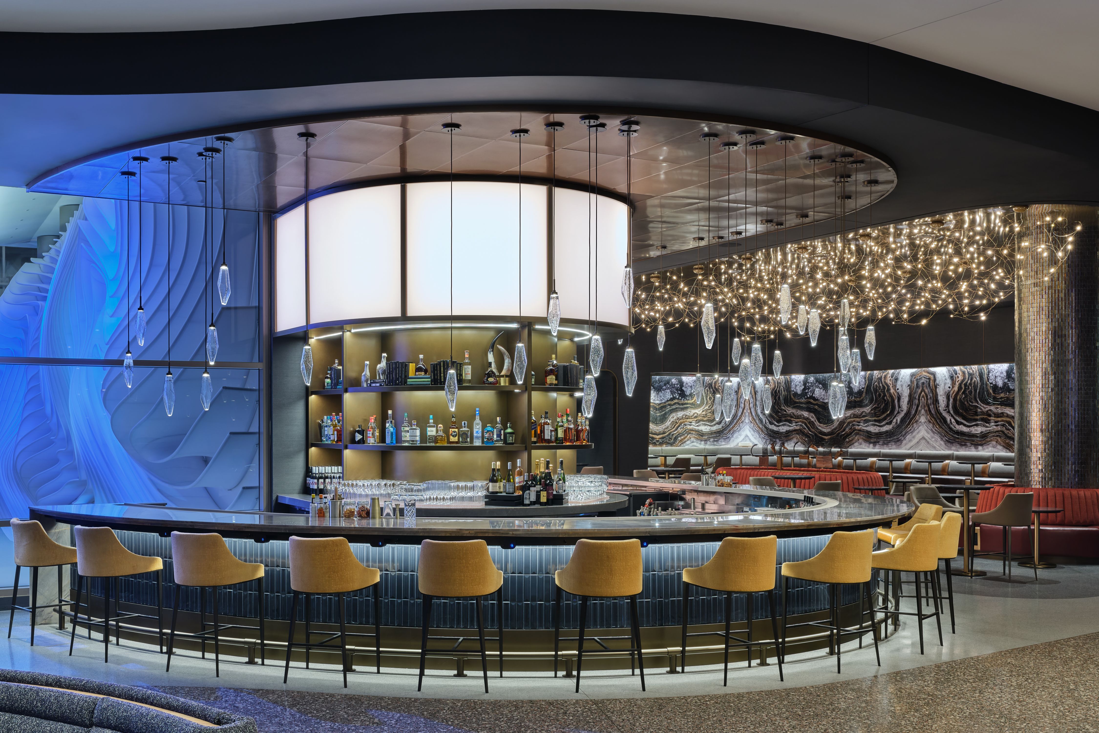 Modern circular bar with yellow stools, crystal pendant lights, illuminated shelves with liquor bottles, and a decorative marble and blue textured wall in the background.