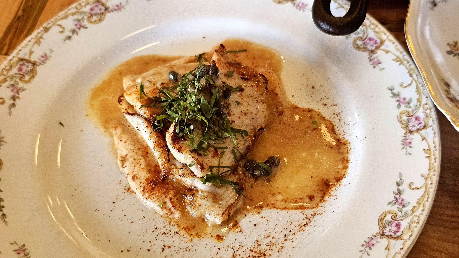 A seared fish fillet sits in a light brown sauce, garnished with chopped herbs and capers, on a decorative floral plate with gold trim.