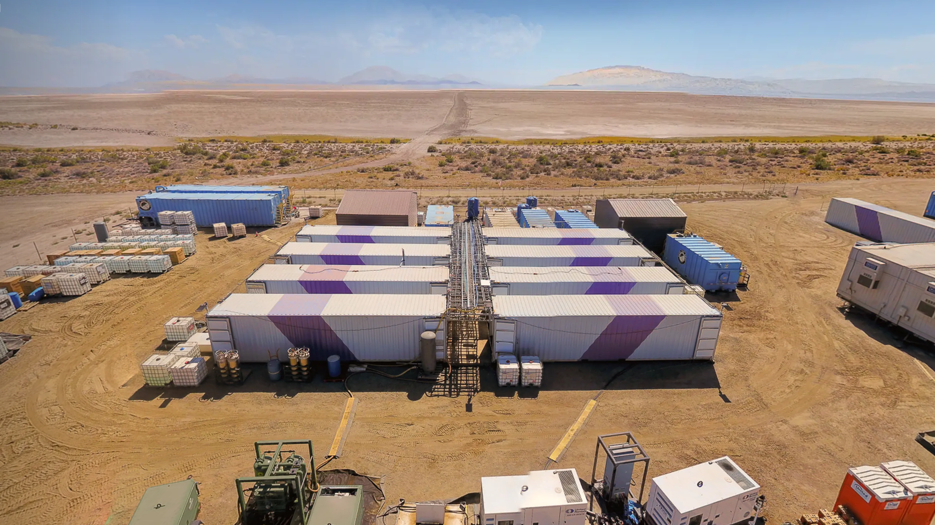 Aerial view of a desert industrial site with white shipping containers painted purple, arranged in rows around a central walkway and equipment, with distant barren plains and mountains.