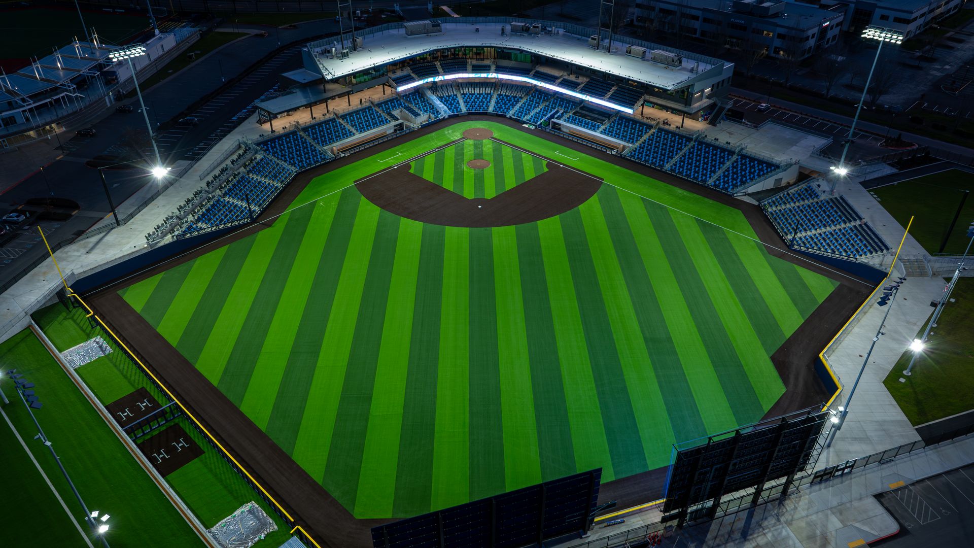 Aerial night view of a baseball stadium: vivid green striped outfield, brown infield dirt, white bases, blue seating, and bright lights illuminating the empty stands.