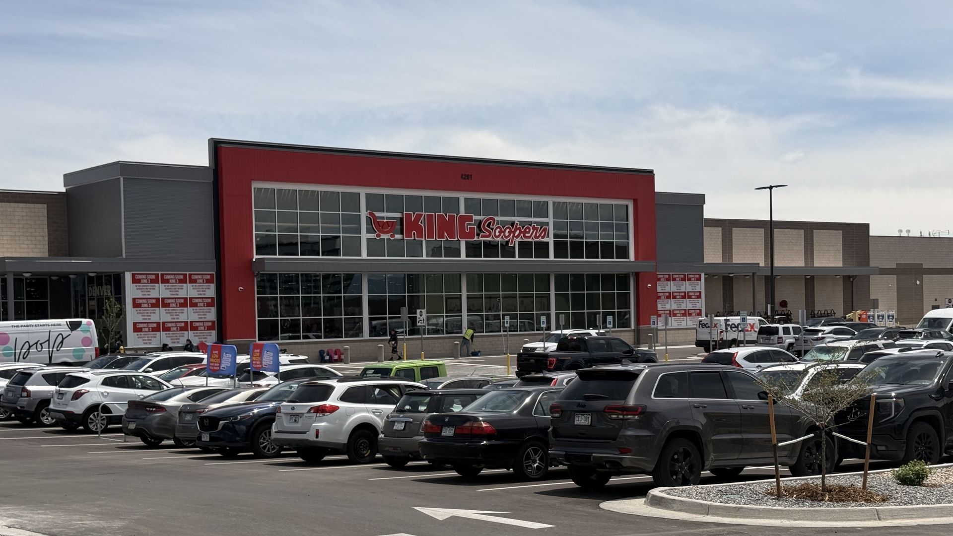 Wide view of a King Soopers supermarket with a bold red facade and large glass front; a crowded parking lot of cars in front, blue sky above with light clouds.