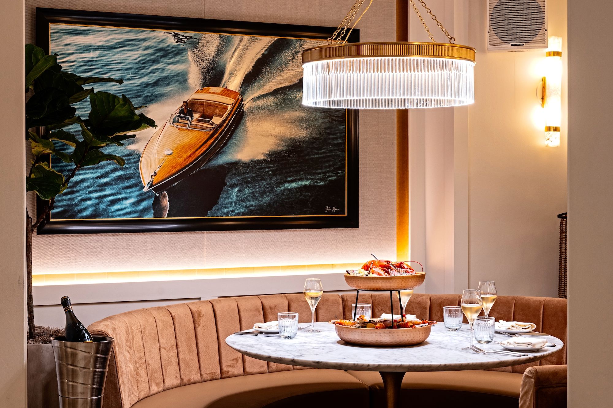 Cozy restaurant corner with round marble table, curved tan velvet seating, seafood served on two-tiered copper trays, champagne glasses, and boat photo on wall under warm lighting.