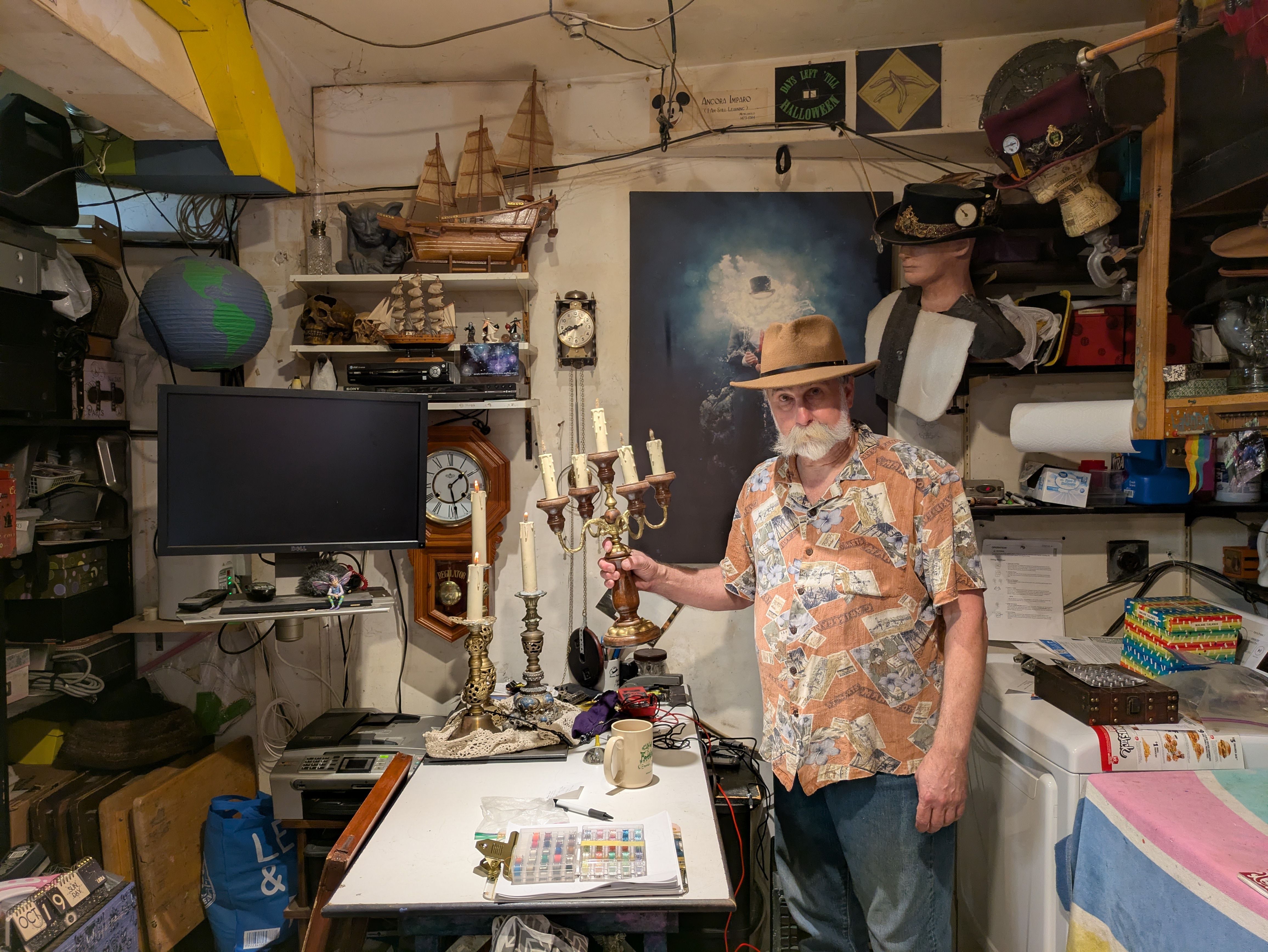 Man with white beard and mustache wearing a tan hat and patterned shirt stands in cluttered room holding a vintage candelabra with candles. Shelves, ship models, and a painting in background.