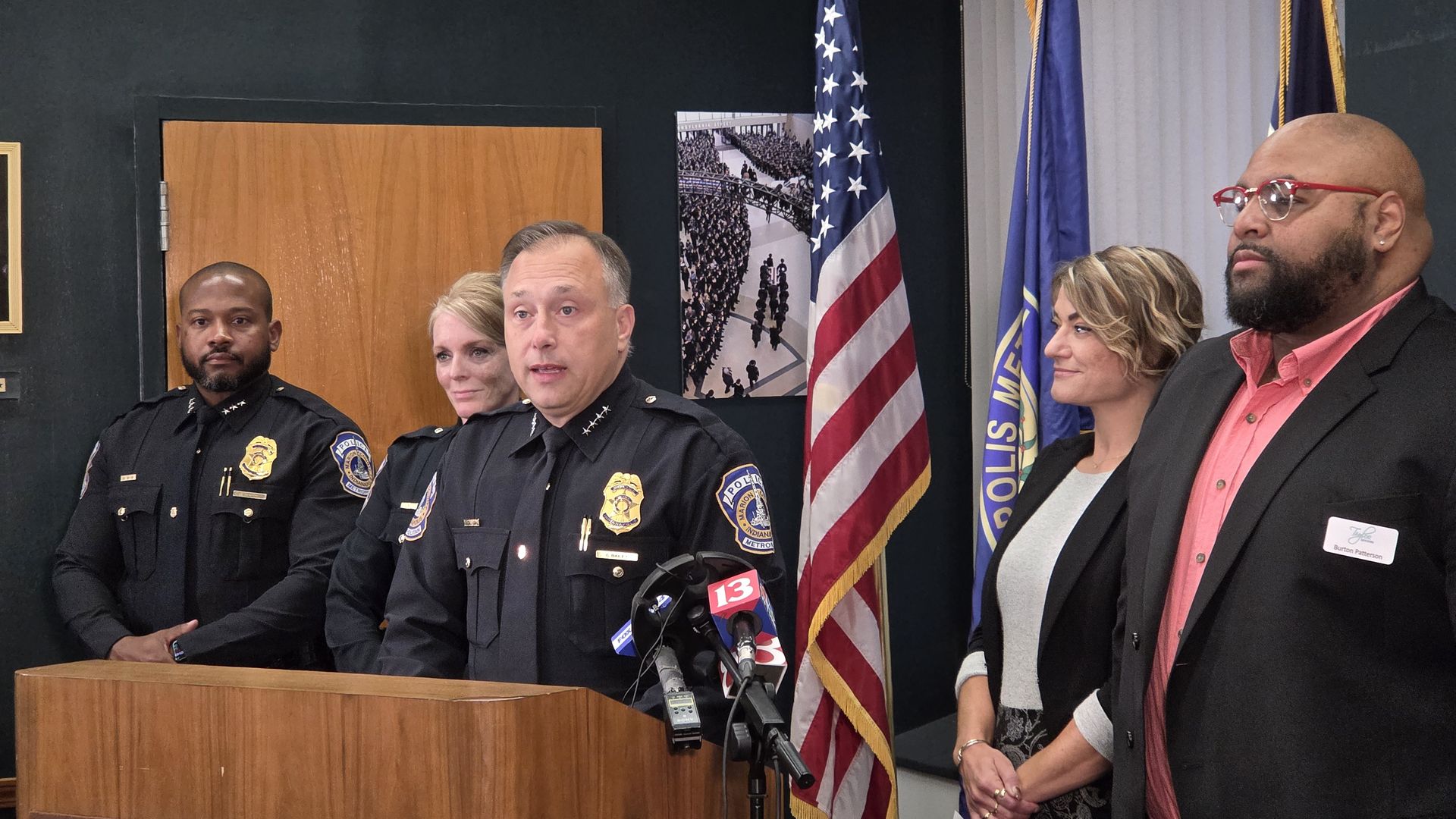 Police chief speaking at podium during press conference, flanked by officers in black uniforms and two formally dressed individuals, with American and police flags in background.