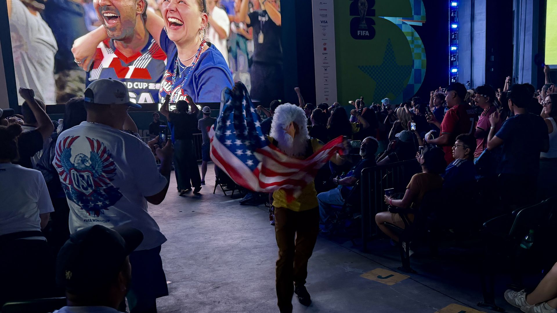A crowded arena watches a large screen showing a jubilant man and woman celebrating; an American flag is waved by fans in the foreground as a live soccer match plays on a side screen.