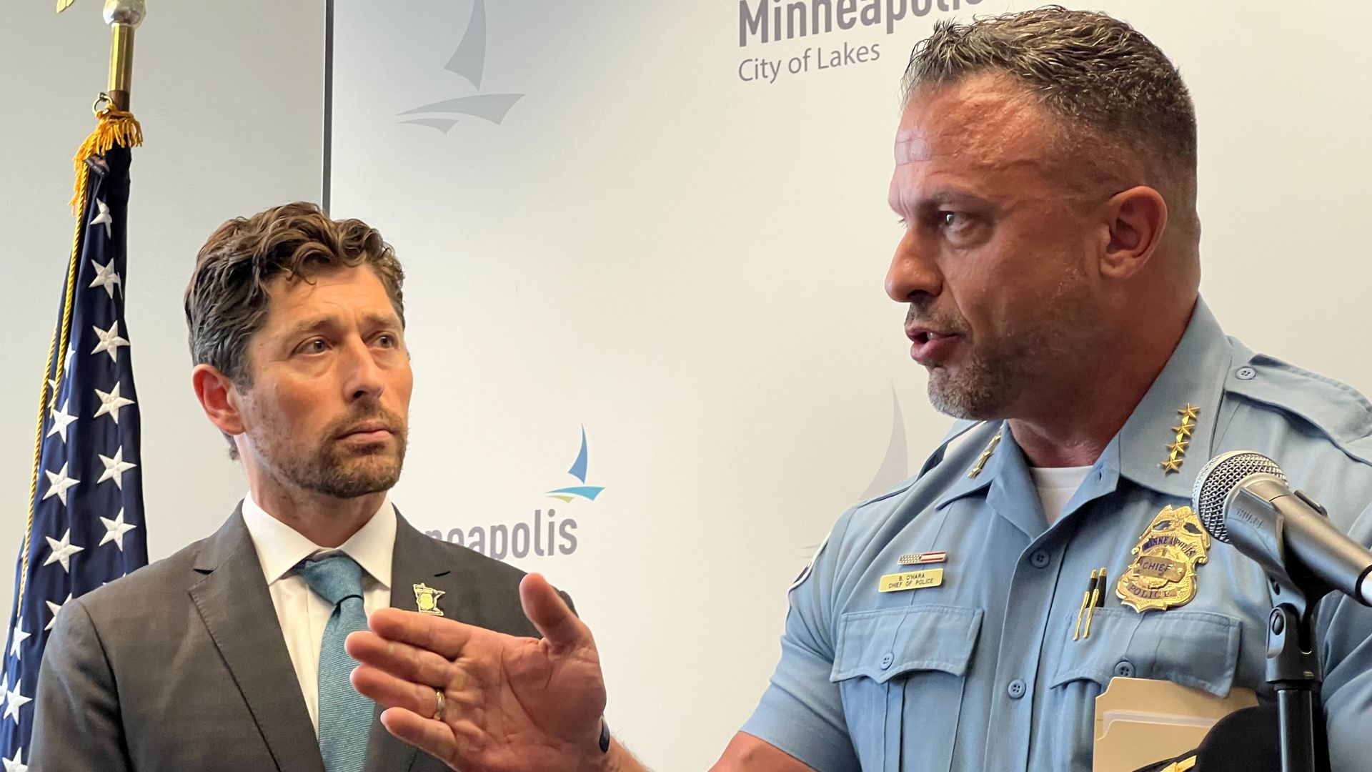 A police chief in a blue uniform with his hat under his arm gestures as another man in a suit looks