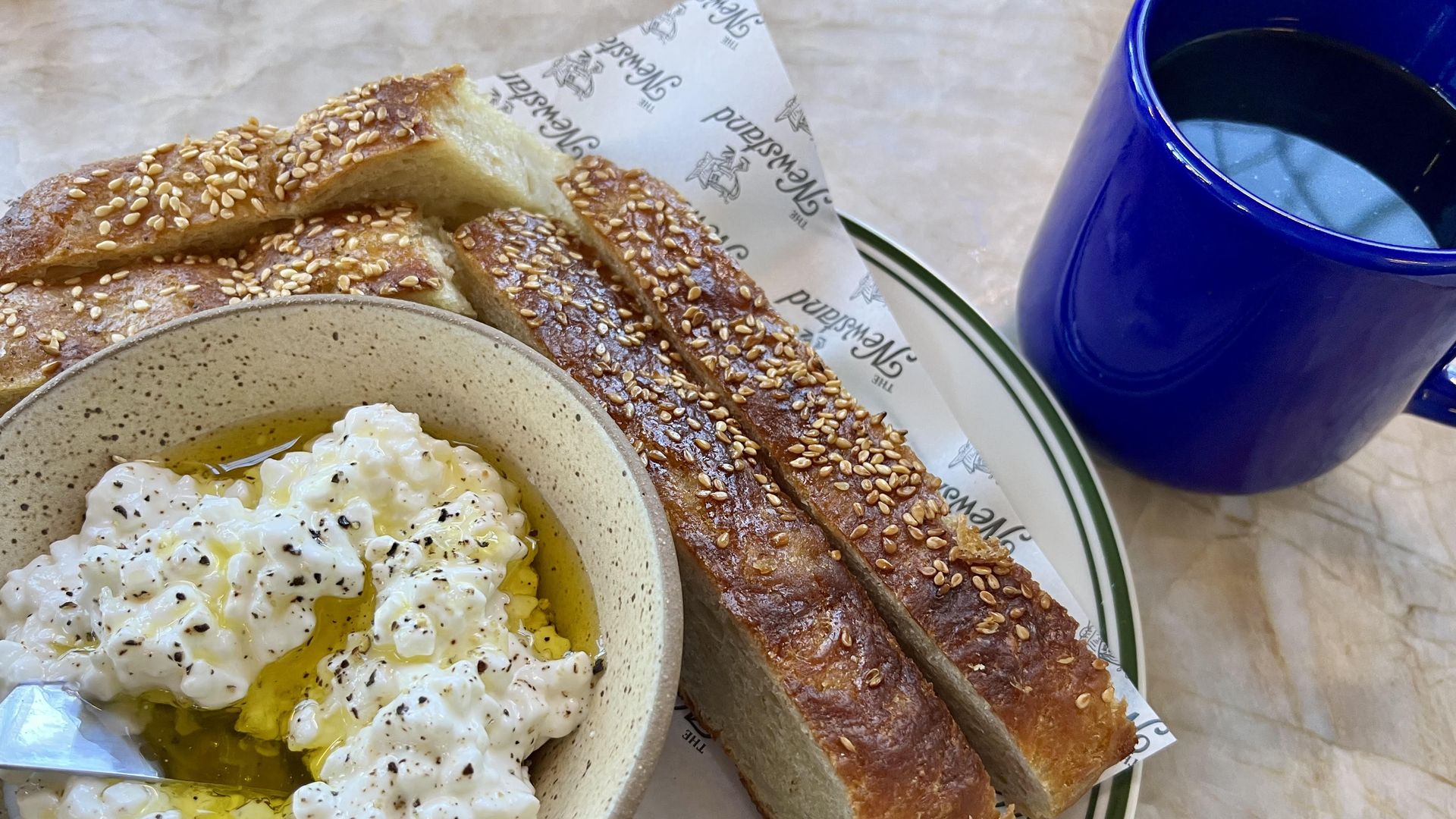 A plate with cottage cheese, covered in olive oil and black pepper, alongside fresh bread and coffee in a blue mug.