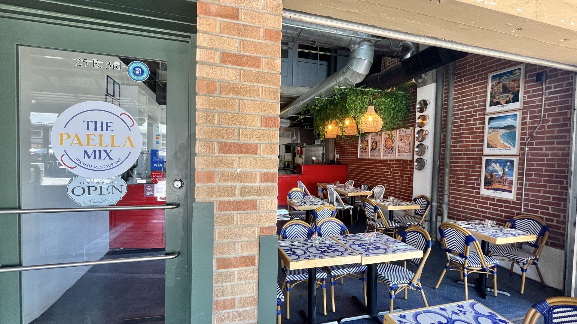 Entry to The Paella Mix Spanish restaurant with open sign. Inside, blue and white tiled tables and wicker chairs, brick walls decorated with Spain city photos, hanging lamps, and green plants.