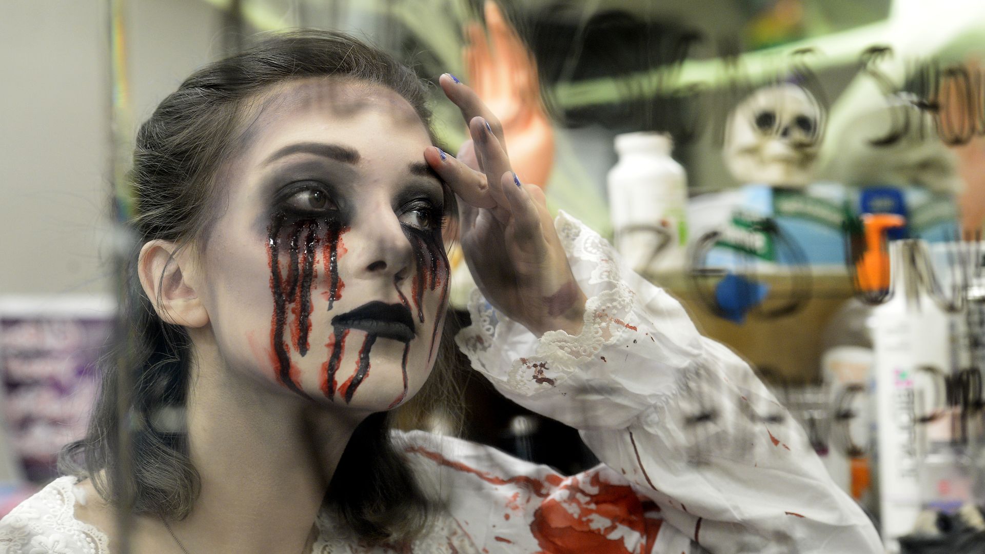 A female high school student with dark eye makeup and red streaks like blood running down her face, wearing a white lace outfit stained with red, posing in front of a mirror with a skull decoration nearby.