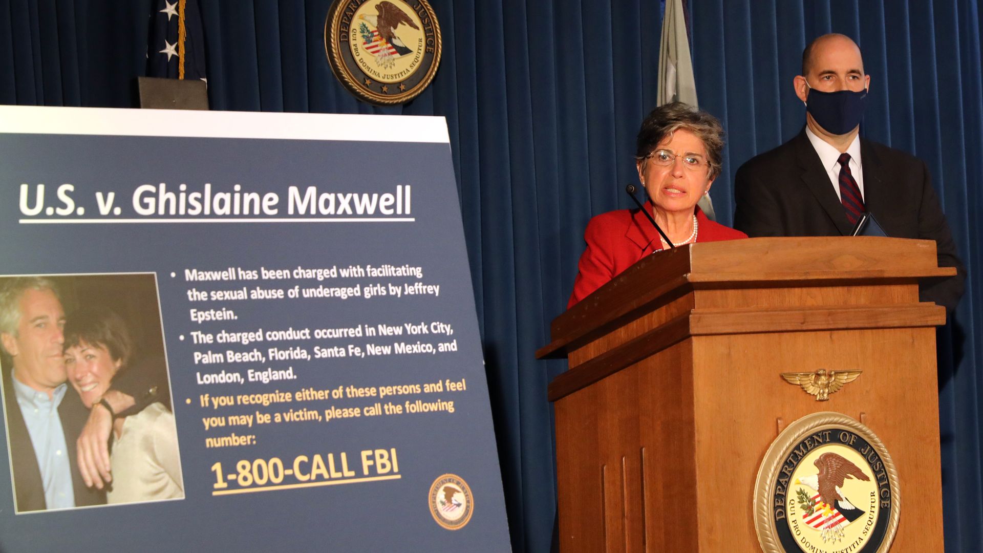 Two officials at a Department of Justice press conference with a large sign detailing charges against Ghislaine Maxwell in the U.S. v. Ghislaine Maxwell case, including contact info for FBI.
