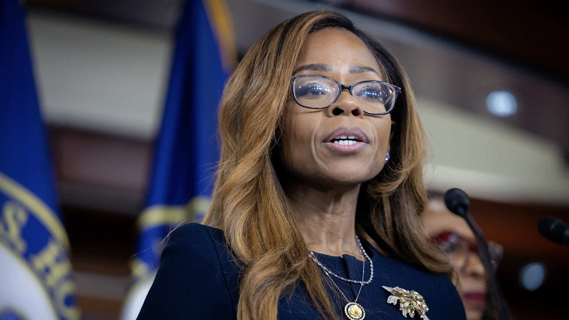 Woman with long hair and glasses speaking at a podium, wearing a navy outfit with a gold brooch and necklace, U.S. House flags in the background, official setting.