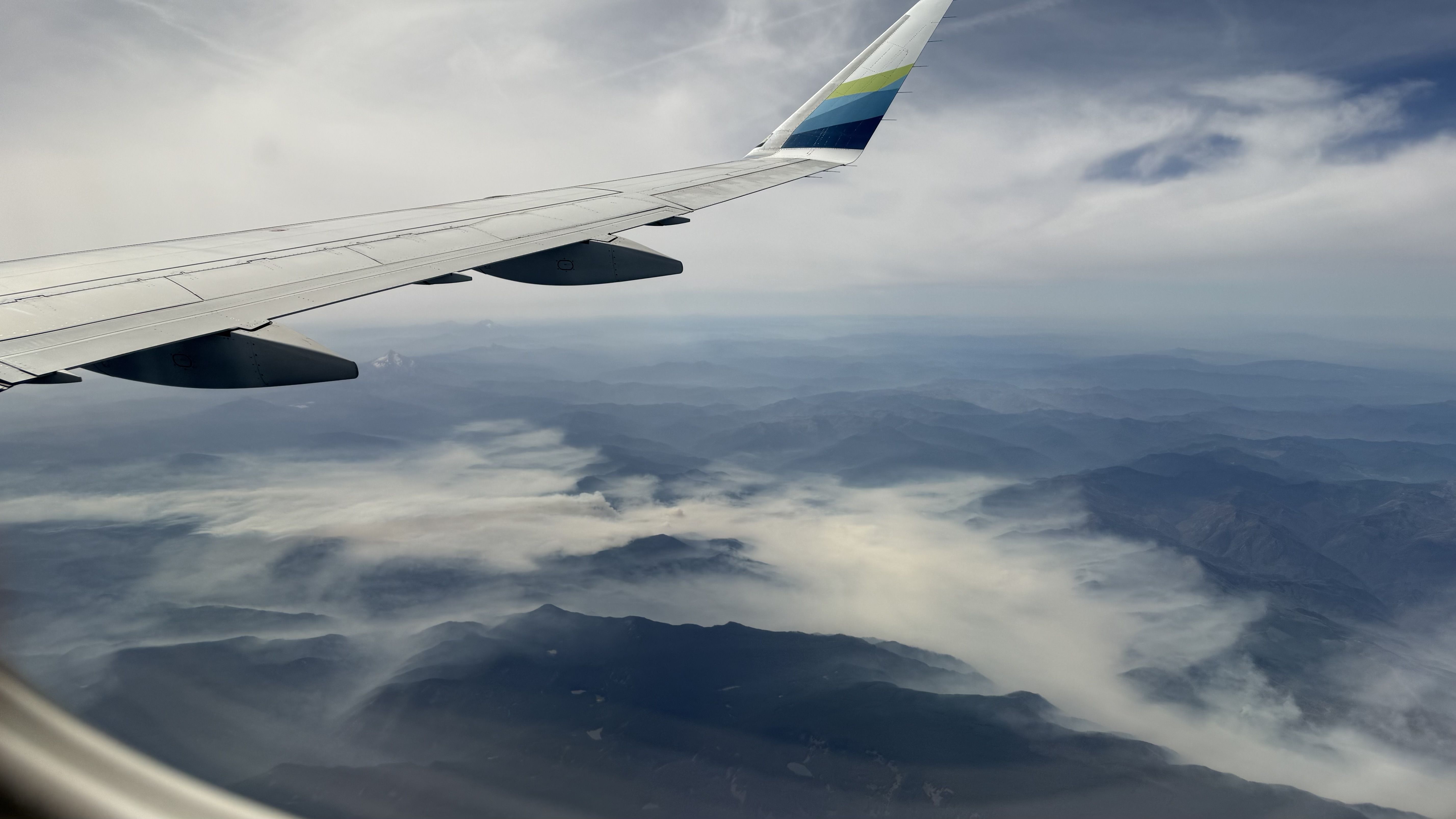 View from a window seat: a white airplane wing with two engine nacelles, a colorful wingtip, and a cloudy sky above a distant mountain range.
