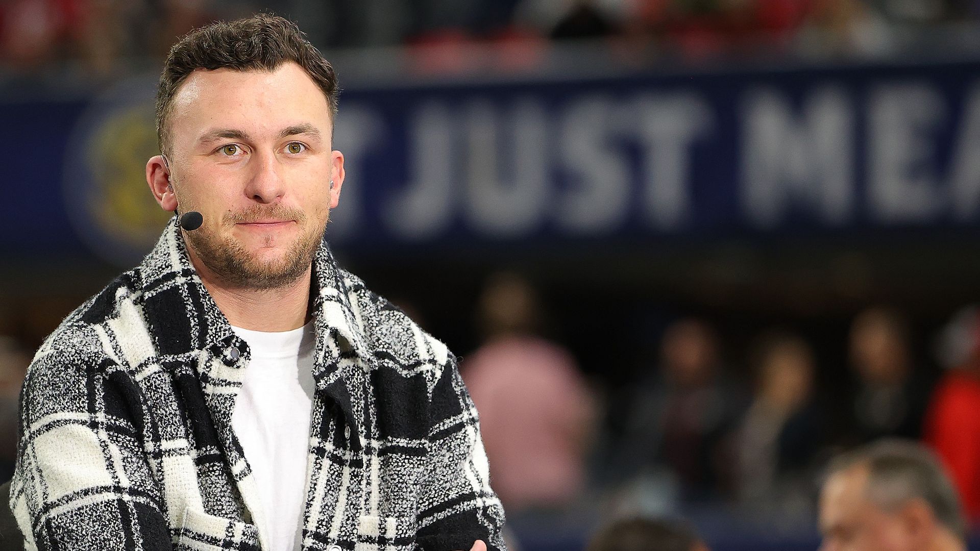 Johnny Manziel, wearing a plaid shirt, sits at a desk at a sporting event with a microphone attached to his head. 
