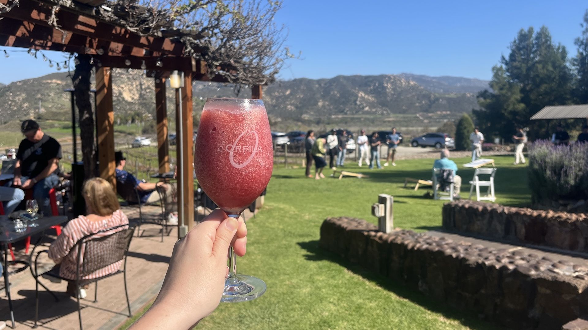 A hand holds up a glass of frozen red sangria at a winery with people playing cornhole and mountains in the background.