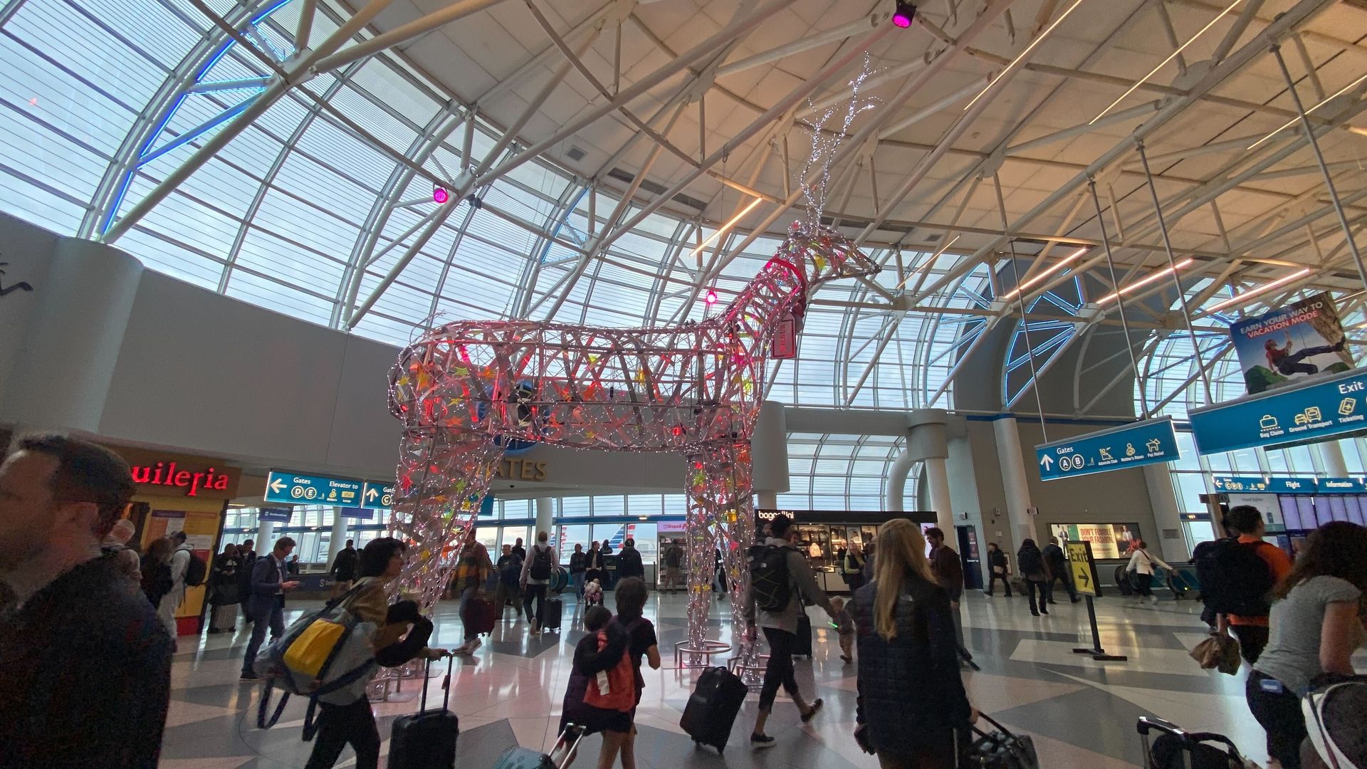 Large illuminated reindeer sculpture with colorful decorations stands in a busy airport terminal with many travelers walking by, pulling suitcases under a translucent curved roof.