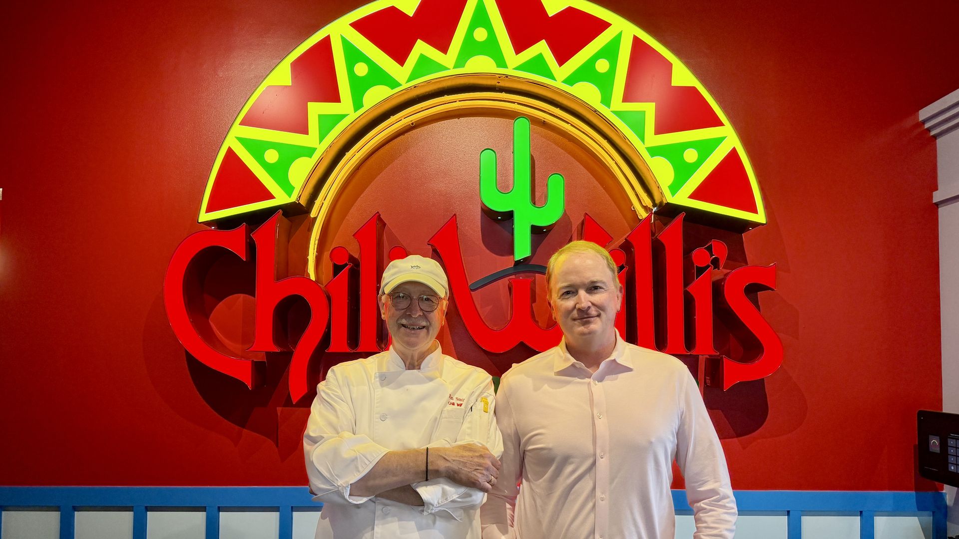 Two men in white chef coats pose before a red wall with a large neon sign: a green cactus emblem inside a yellow-green border, and red lettering partially visible behind them.
