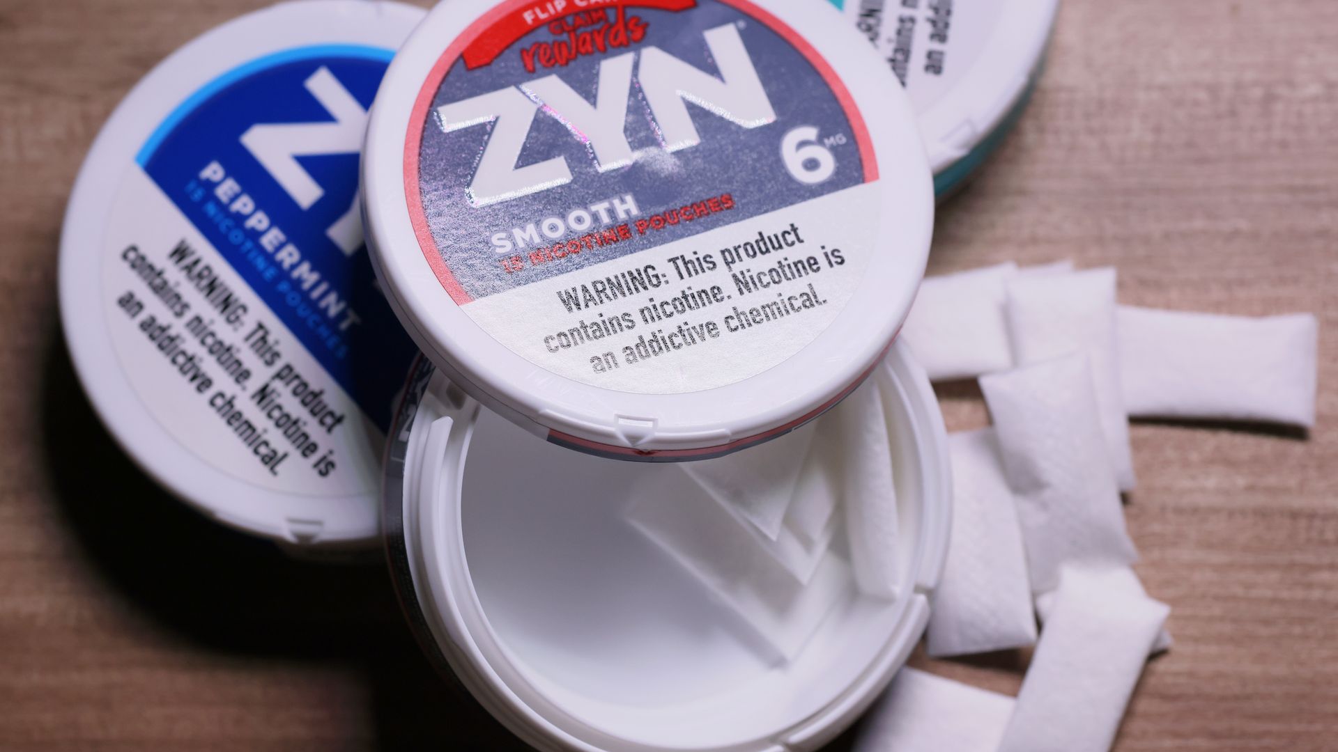 Open can of ZYN Smooth 6 mg nicotine pouches with some pouches spilled beside it, and another closed can of ZYN Peppermint on a wooden surface.