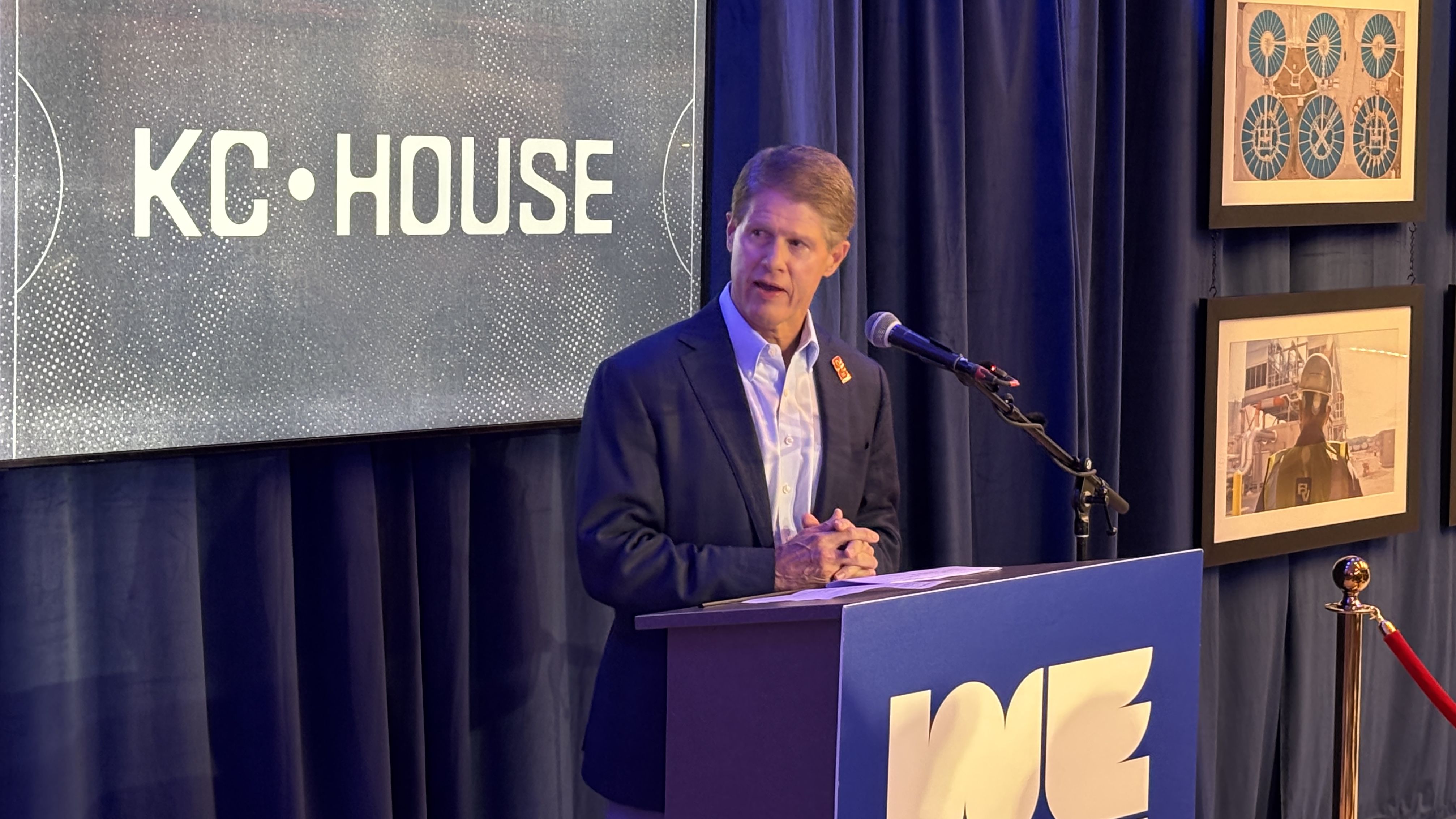 Man in a dark suit speaks at a blue podium with a microphone; behind him a screen reads "KC HOUSE," blue curtains, and framed wall art.