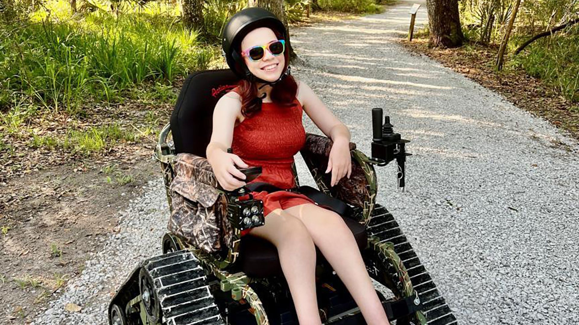 Photo shows a young woman in a track wheelchair on a gravel path. The chair is covered in a camo print and has a triangle-shaped track instead of wheels. She is wearing a helmet and sunglasses and using hand controls to move the chair.