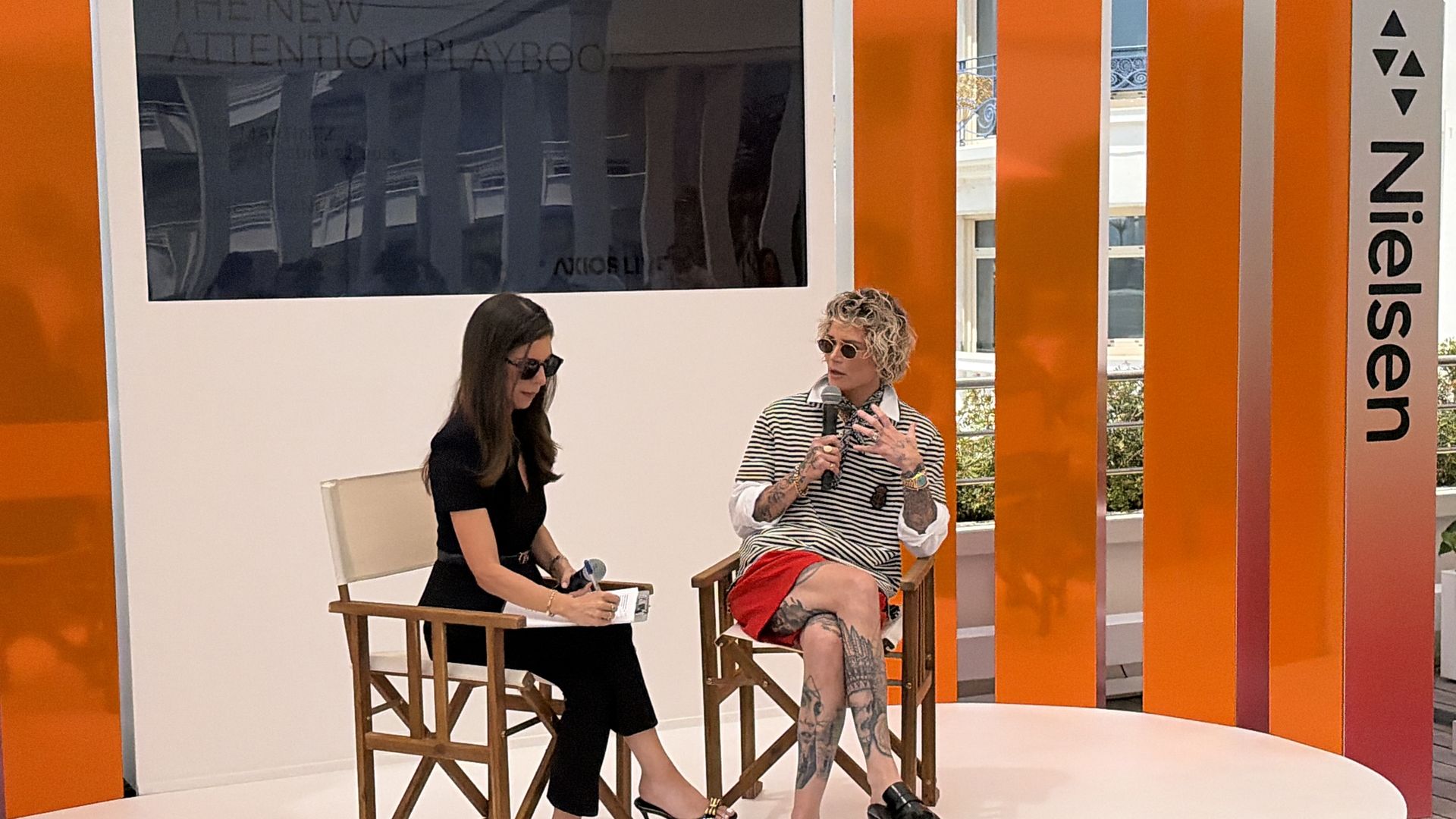 Two women sit on a white stage in director chairs among tall orange panels; one writes in a notebook, the other speaks into a microphone. A large screen and the vertical word "Universe" are behind them.