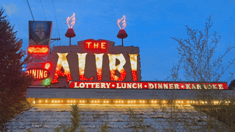 Colorful neon signage at dusk for "The Alibi" with lit flames, advertising lottery, lunch, dinner, and karaoke, against a blue sky and surrounded by trees.