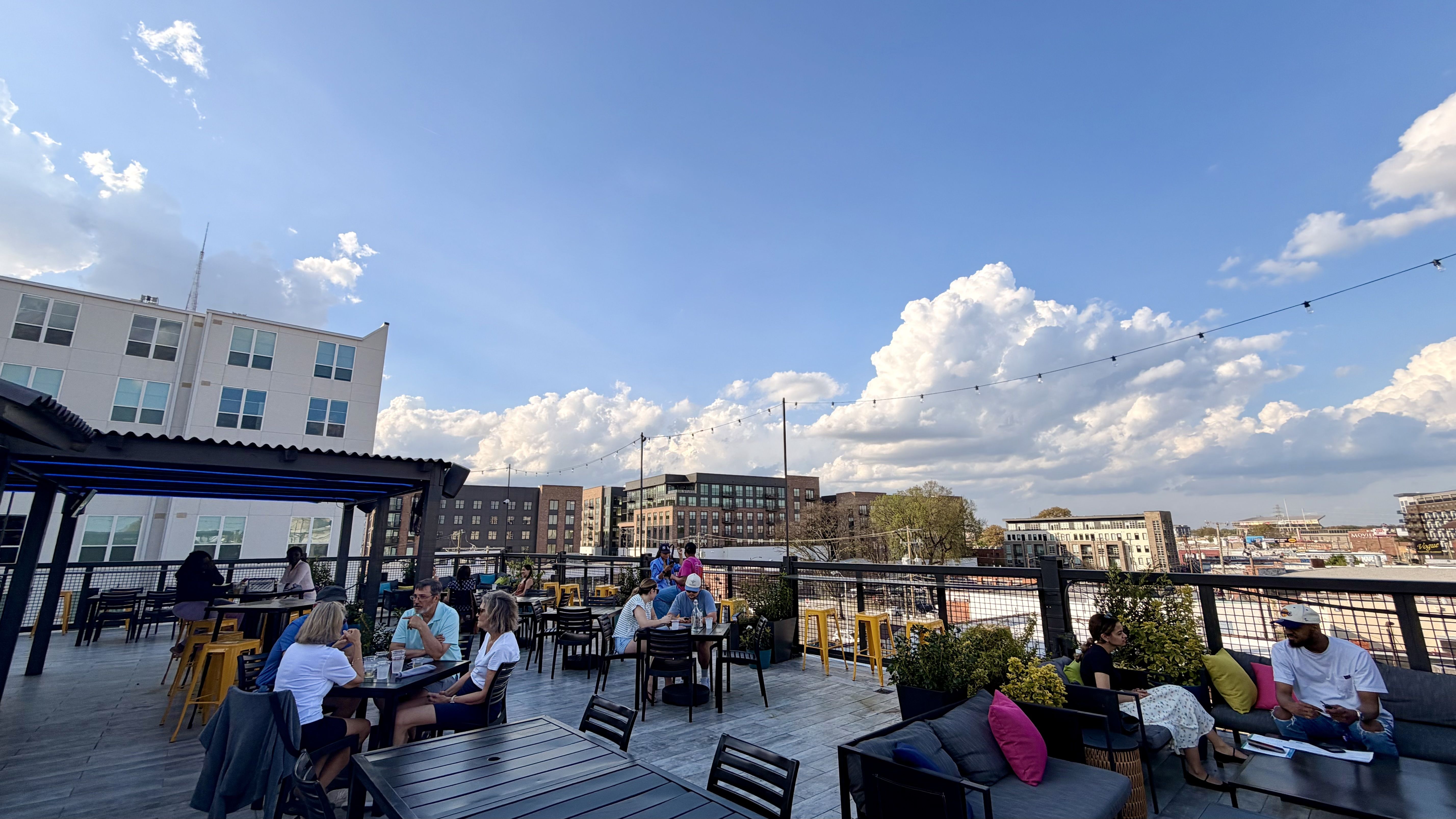 Rooftop cafe scene on a sunny day: people seated at black tables with yellow stools, string lights overhead, modern buildings and city skyline in the distance, blue sky with fluffy clouds.