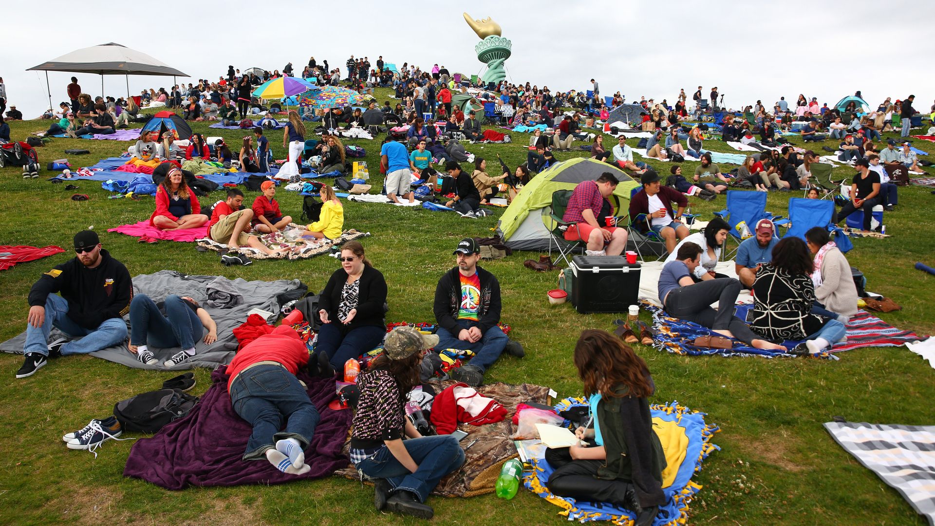 People sit on a hillside with an inflated hand of the Statue of Liberty in the background.