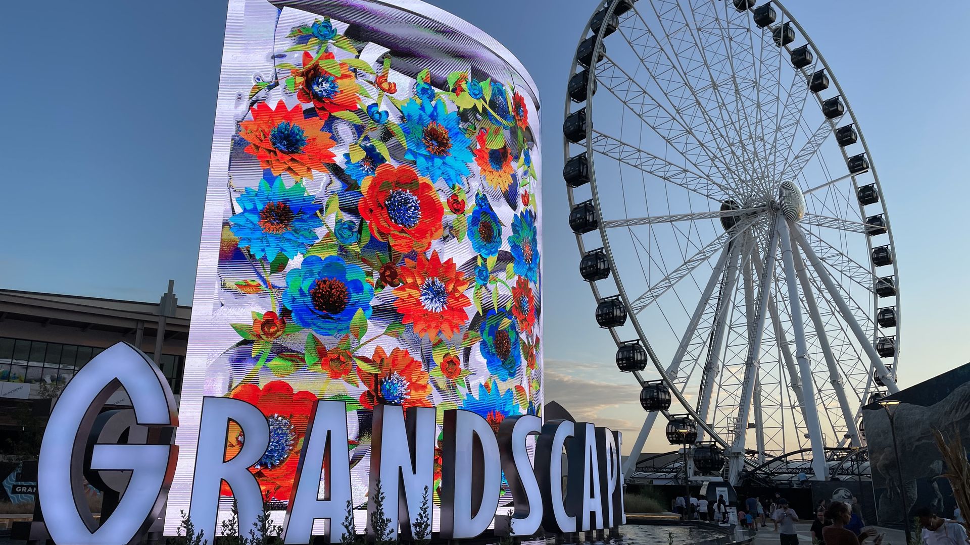 An LED sign is illuminated with a floral pattern in front of a tall Ferris wheel at sunset.