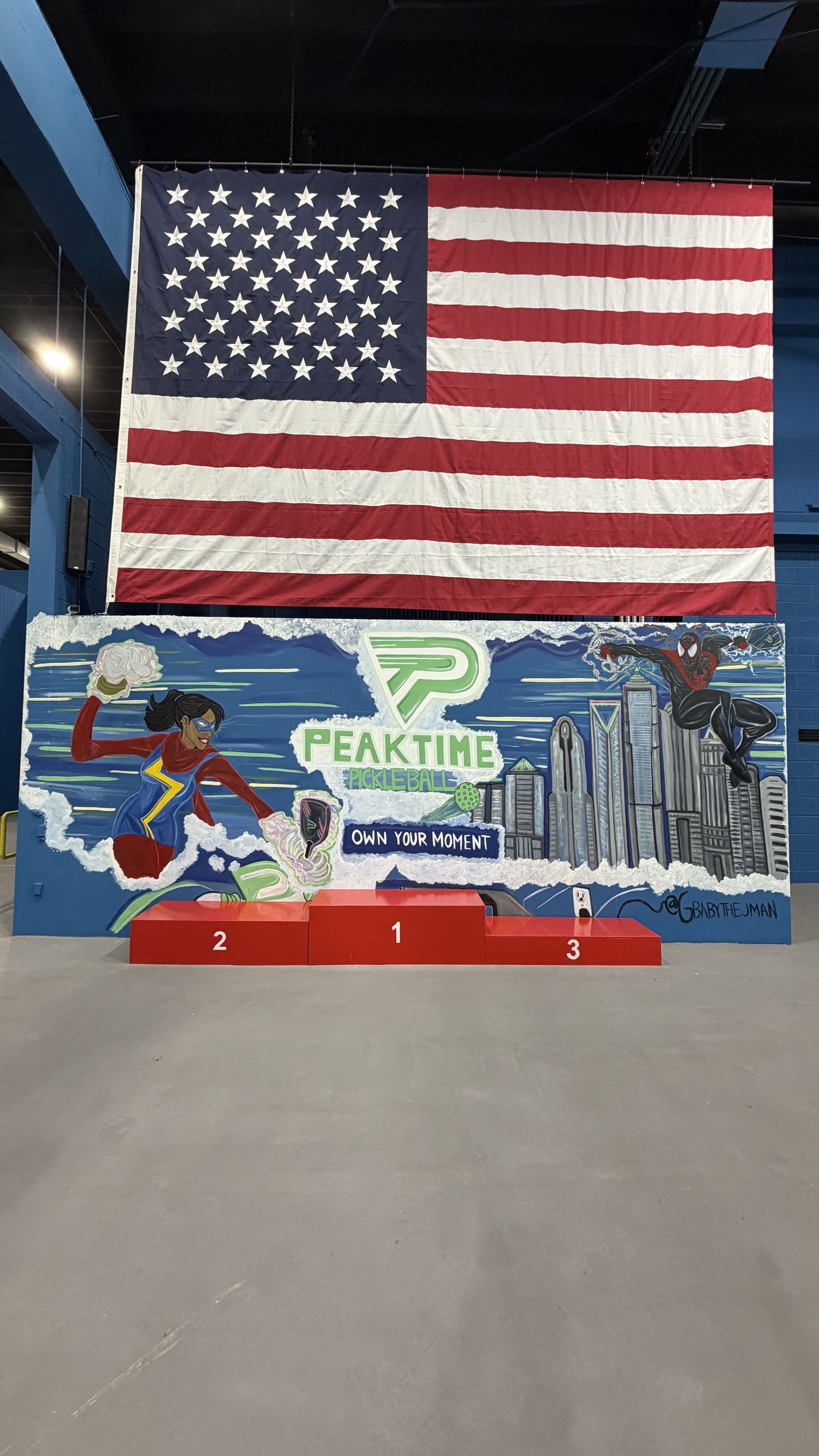 Inside a venue, a large American flag hangs above a vibrant mural for "PeakTime Pickleball" featuring superheroes, a city skyline, and red podiums labeled 1, 2, 3.