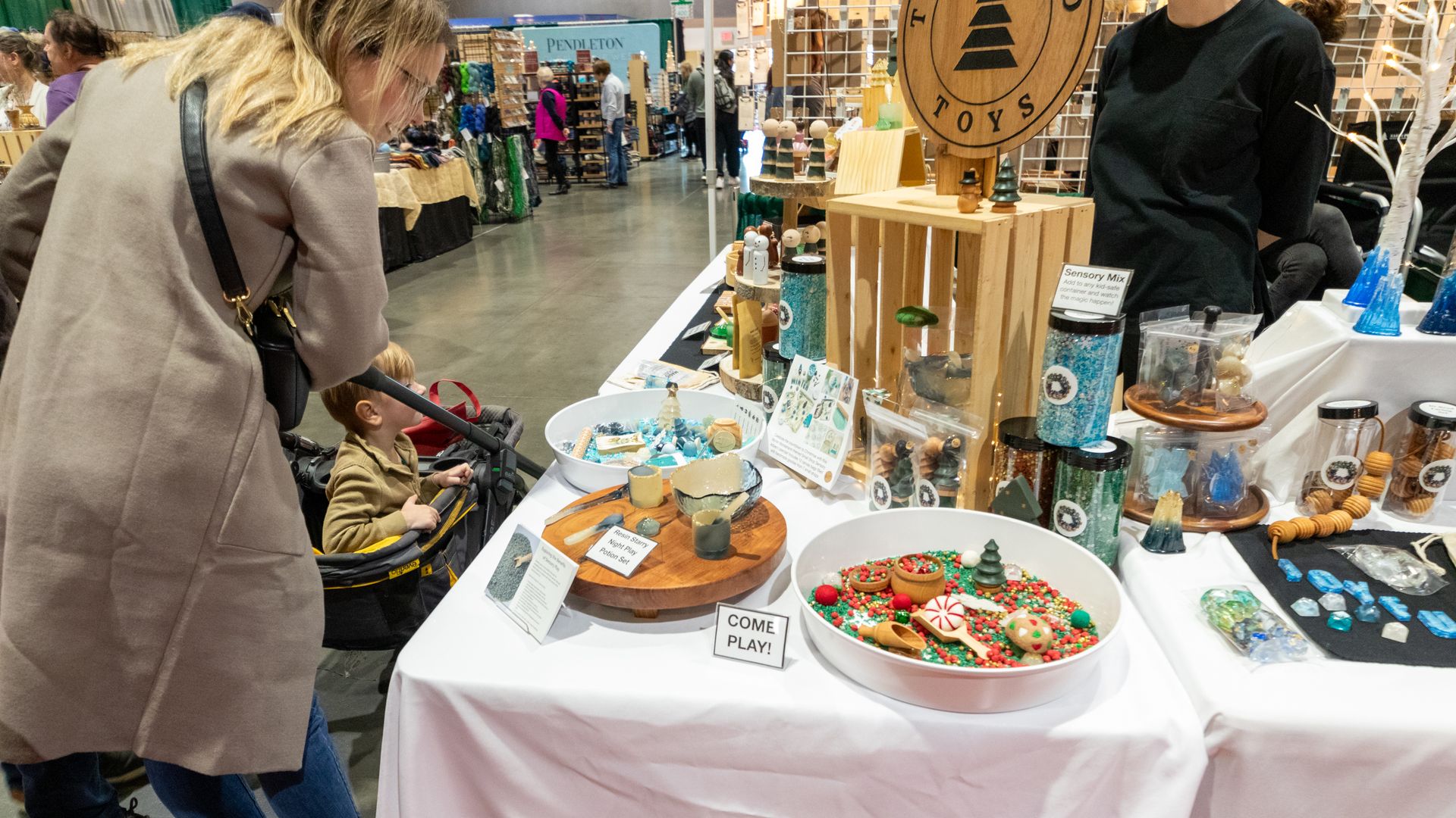 A woman leans over a stroller with a child at a toy booth with wooden toys, colorful sensory bins, and decorative items at an indoor market or fair.