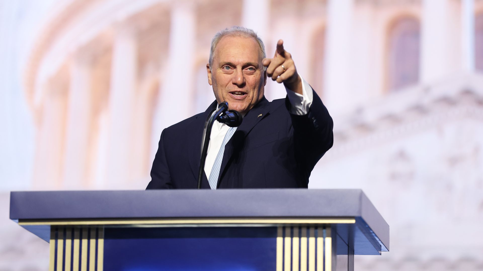 A man in a navy suit at a podium, pointing toward the audience, speaking into a microphone. Behind him are large, blurred beige arches and columns; the stage is blue with gold accents.