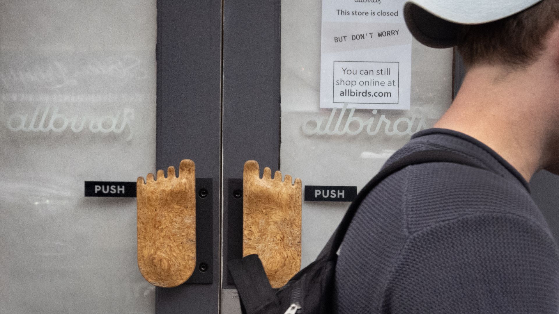 Glass storefront doors with black PUSH signs and two tall, wooden finger-shaped handles; a person in a cap and backpack stands to the right, showing a partial profile.