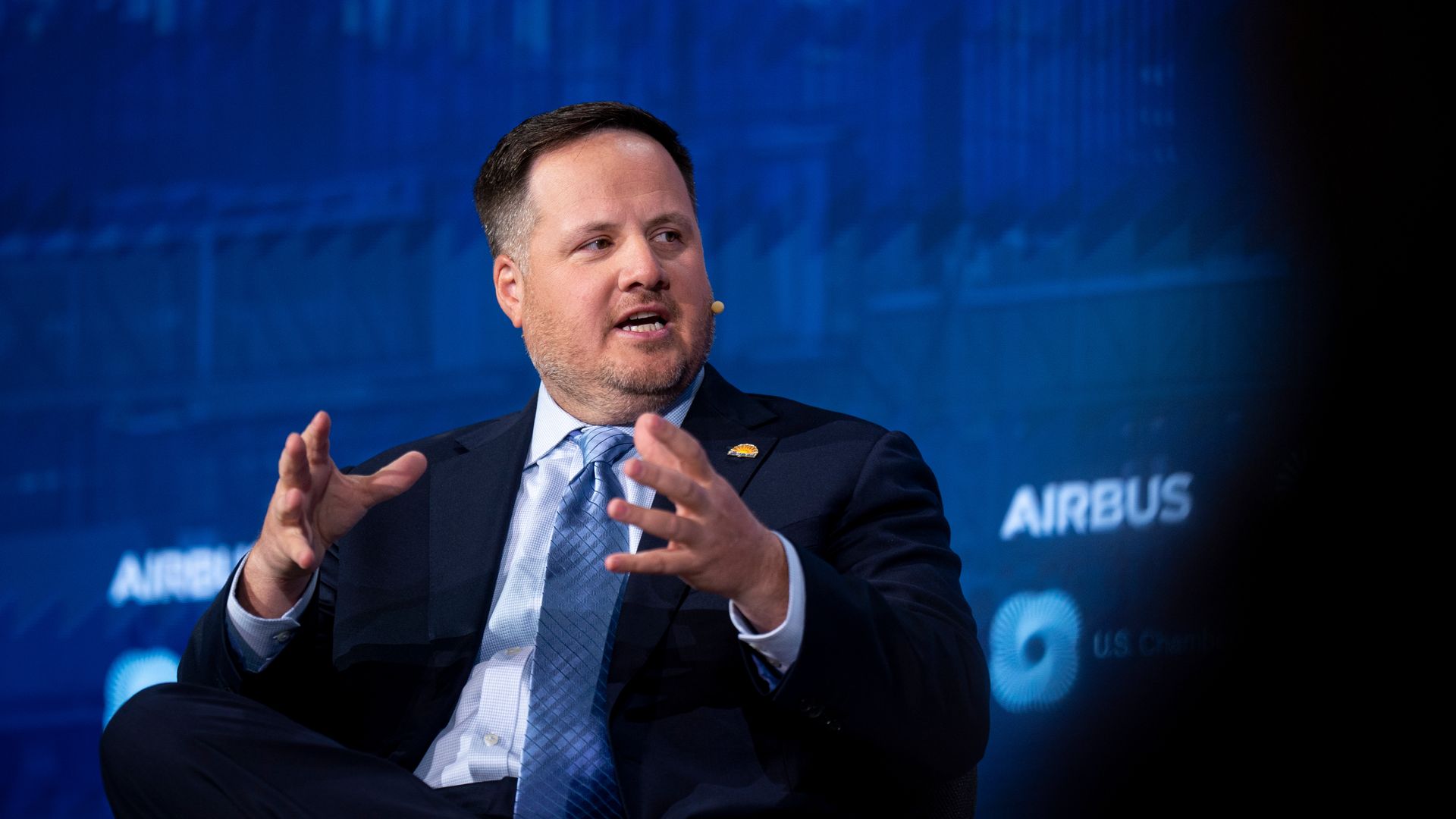 A man in a dark suit and blue tie speaks on a stage with a blue backdrop, gesturing with his hands; a small lapel pin is visible and Airbus branding can be seen in the background.