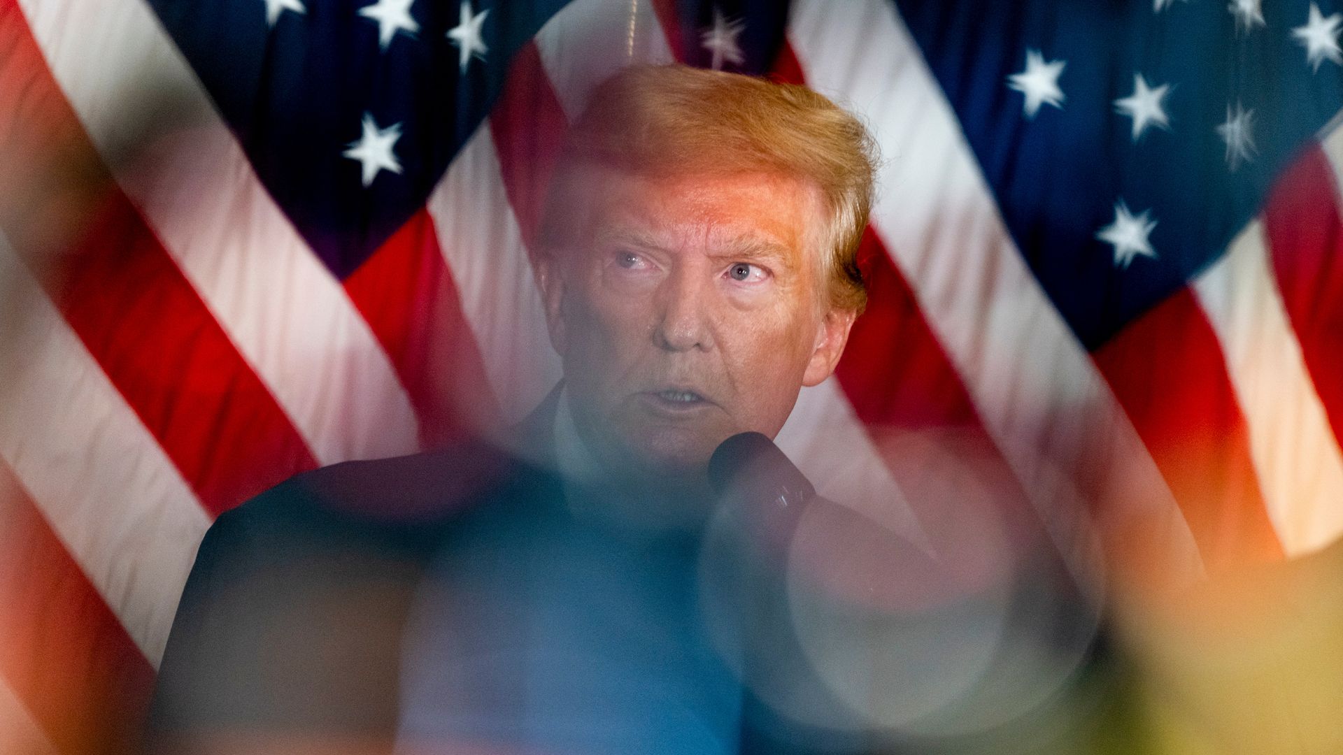 Donald Trump, wearing a dark gray suit standing in front of American flags.