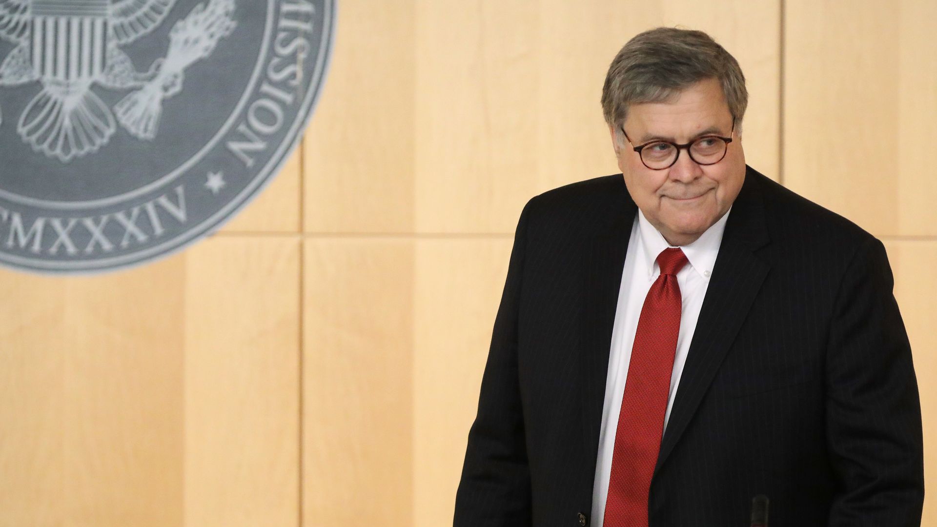 In this image, Barr smiles and walks while wearing a suit and tie