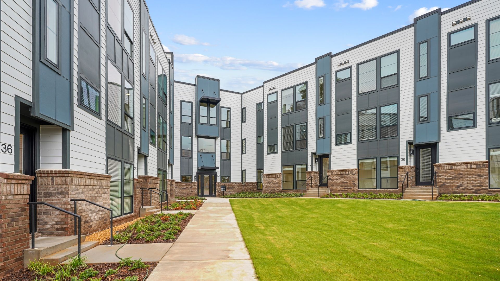 Modern apartment complex with white and gray siding, brick accents, large windows, a green lawn, flower beds and a clear blue sky overhead.
