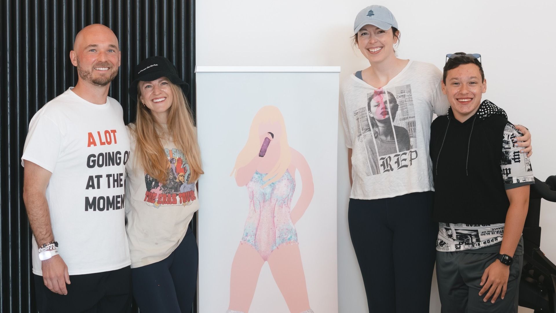Four smiling people standing next to a poster of a blonde woman holding a microphone. The man on left wears a white shirt with red and black text; two women wear graphic tees and caps; a boy in black and white music-print hoodie.