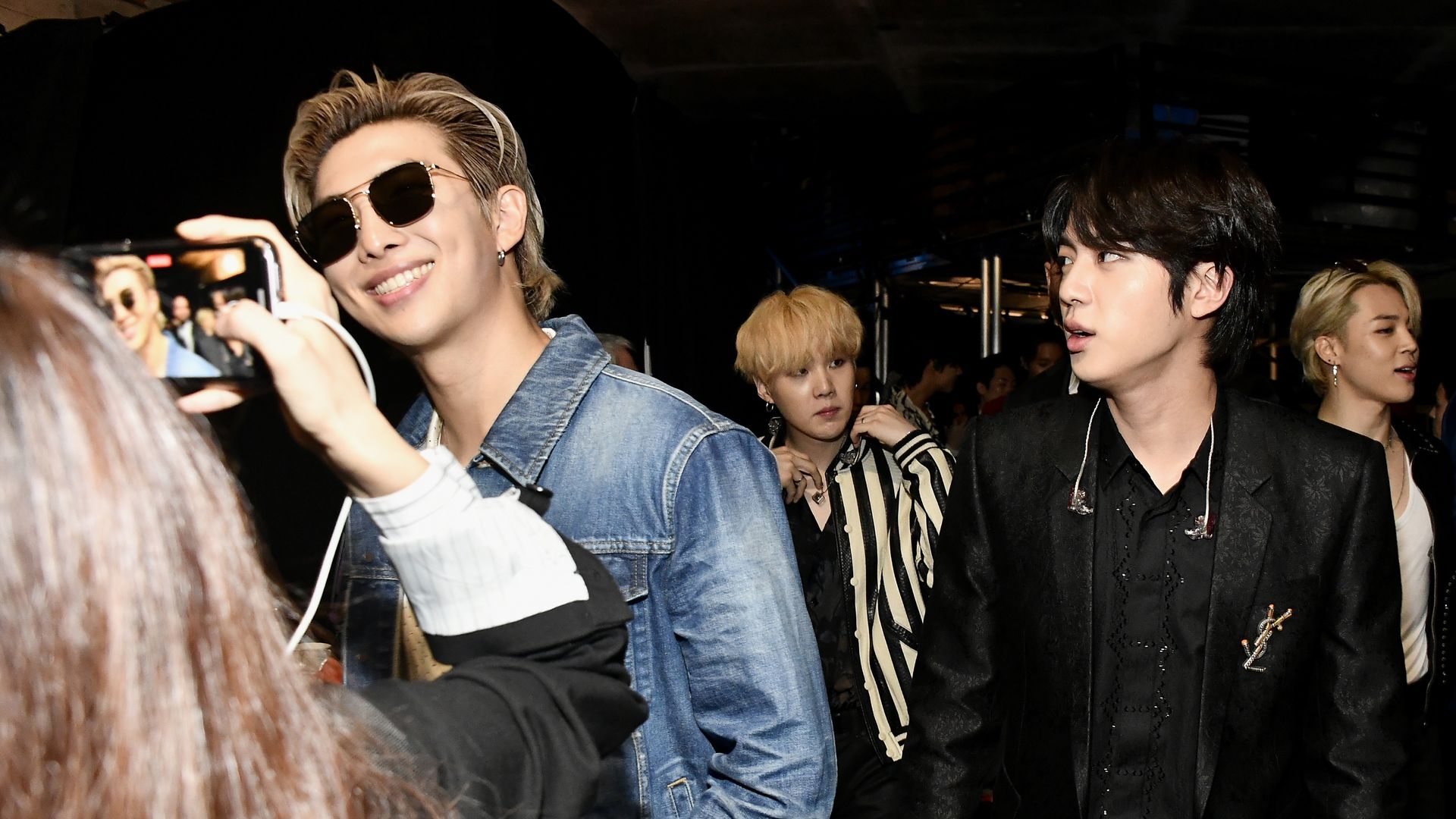 Four men in stylish outfits, one in a blue denim jacket and sunglasses smiling at a phone camera, another in a black suit with ear monitors, and two in striped and black clothing, indoors with dark background.