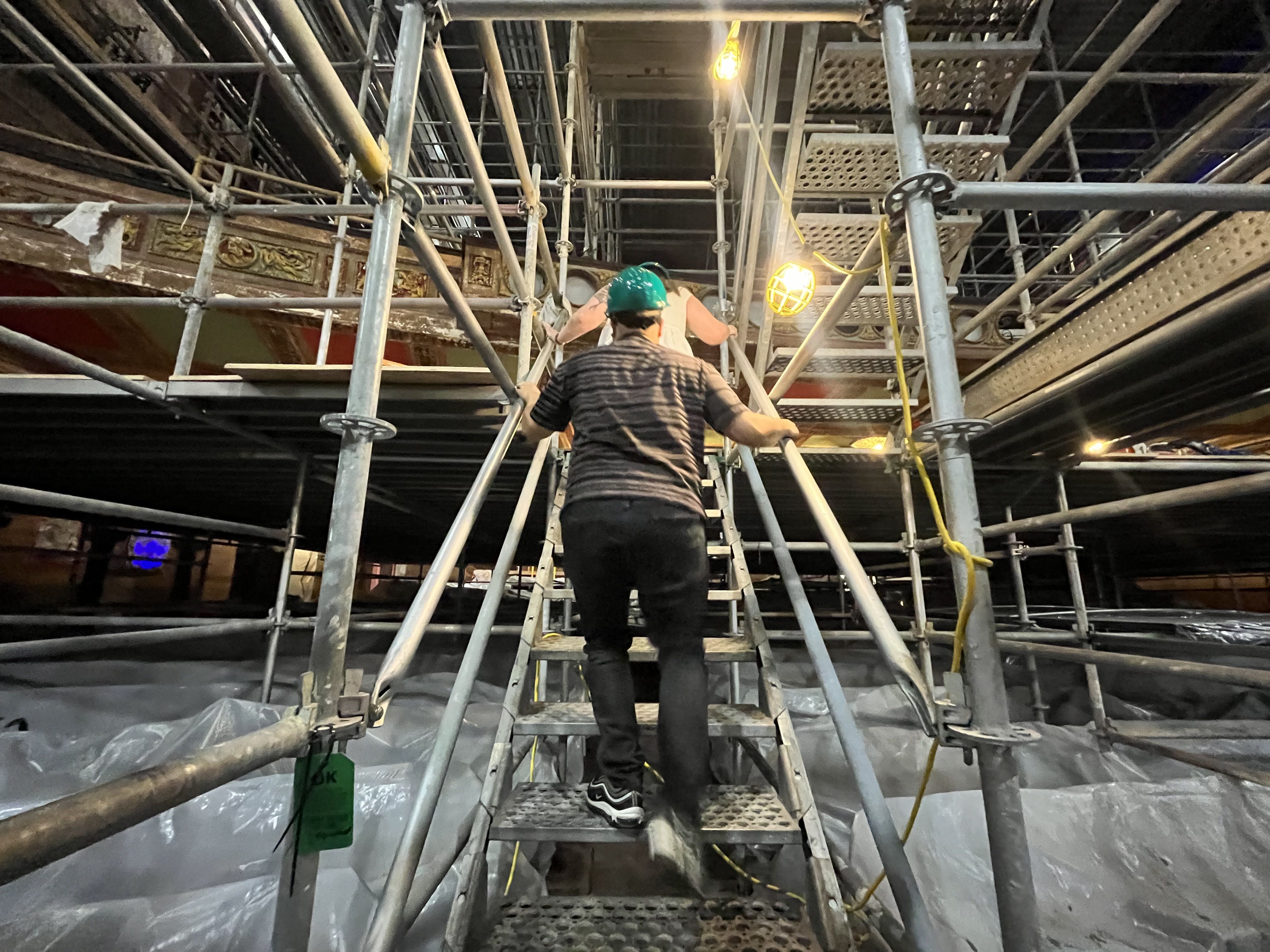 Man in a green hard hat climbs a metal scaffold staircase amid dense scaffolding, pipes, and hanging yellow lights inside a dim construction site.
