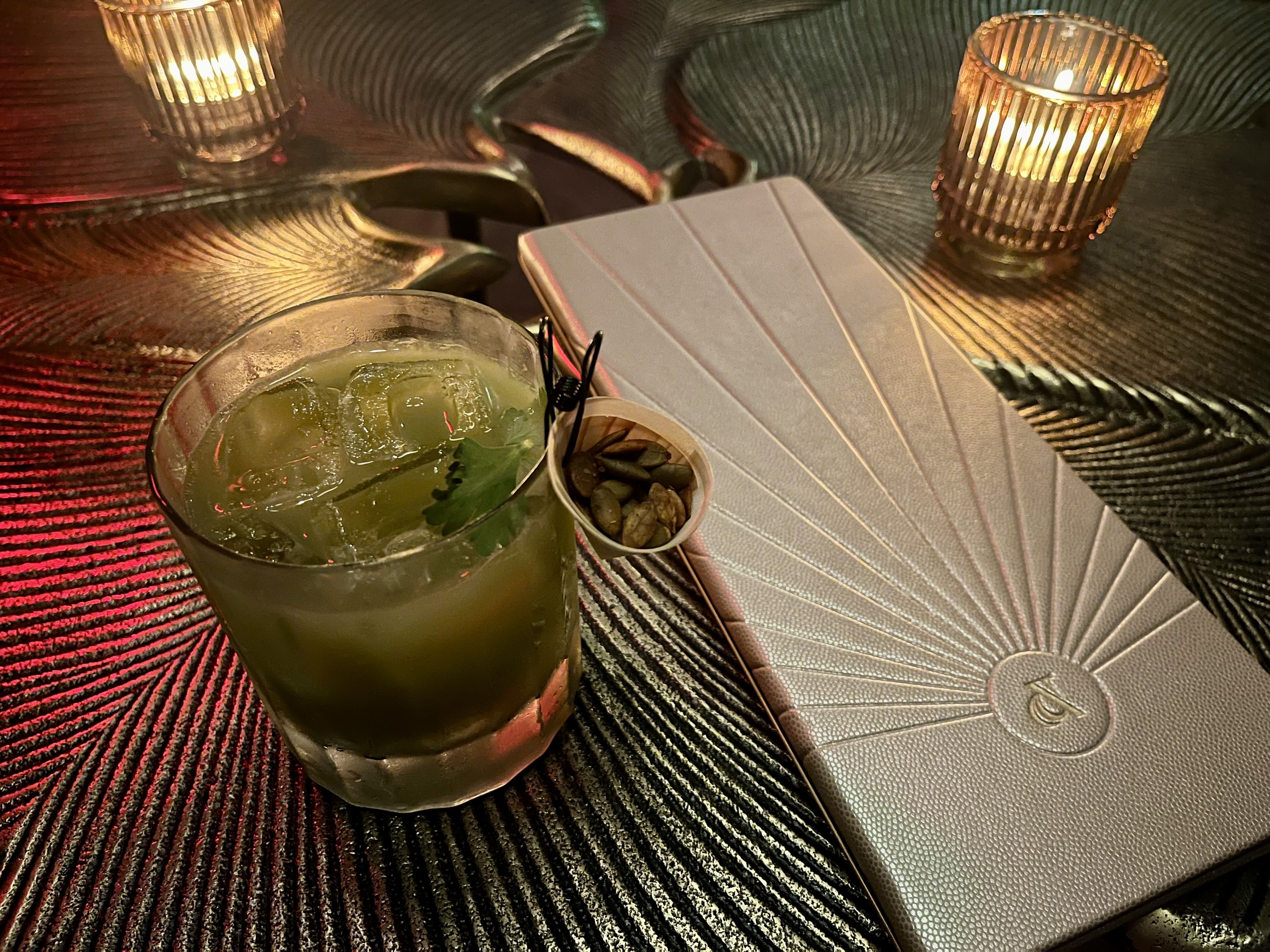 A green drink with ice and garnished with cilantro and a packet of pumpkin seeds clipped to the crystal glass. To the right of the drink, a silver menu embossed with an ampersand.