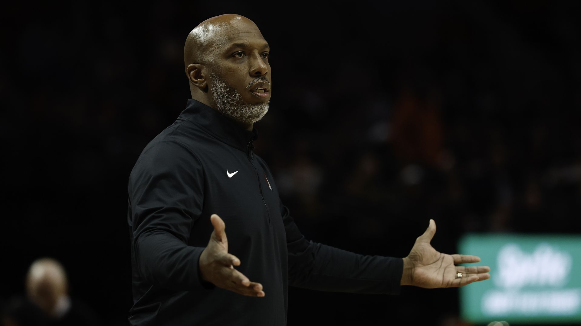 Portland head coach Chauncey Billups wearing a black Nike jacket, standing with arms outstretched, appearing to explain or question something in a dimly lit indoor setting.